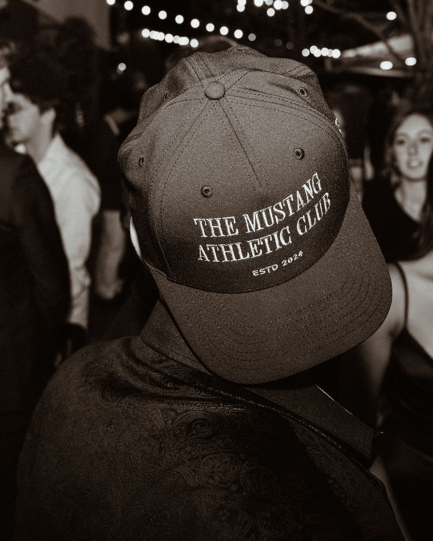 Black and white photo of person wearing The Mustang Athletic Club ESTD 2024 snapback cap at event.