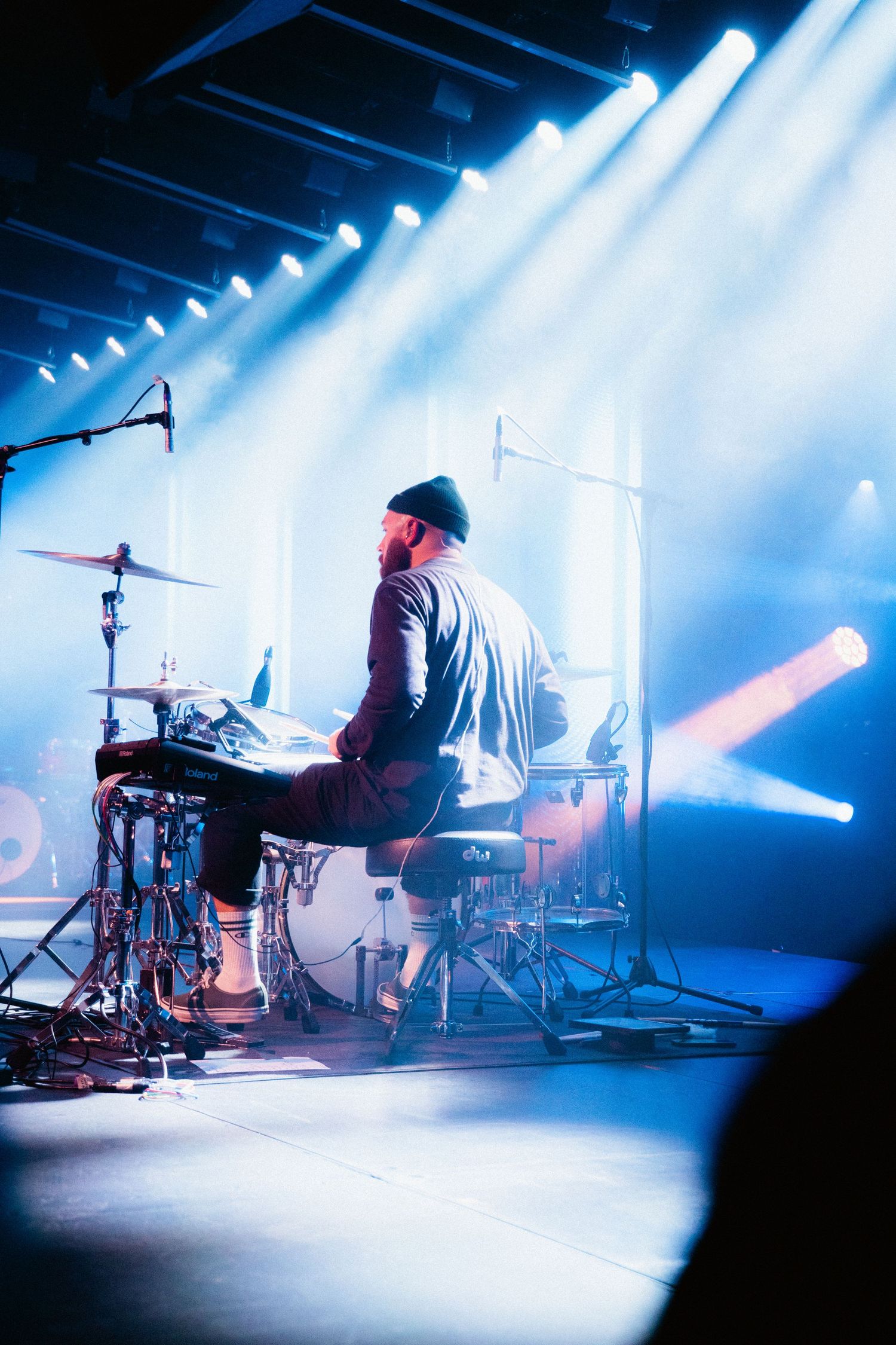 Bearded drummer seen from behind playing drums under blue and white concert stage lights.