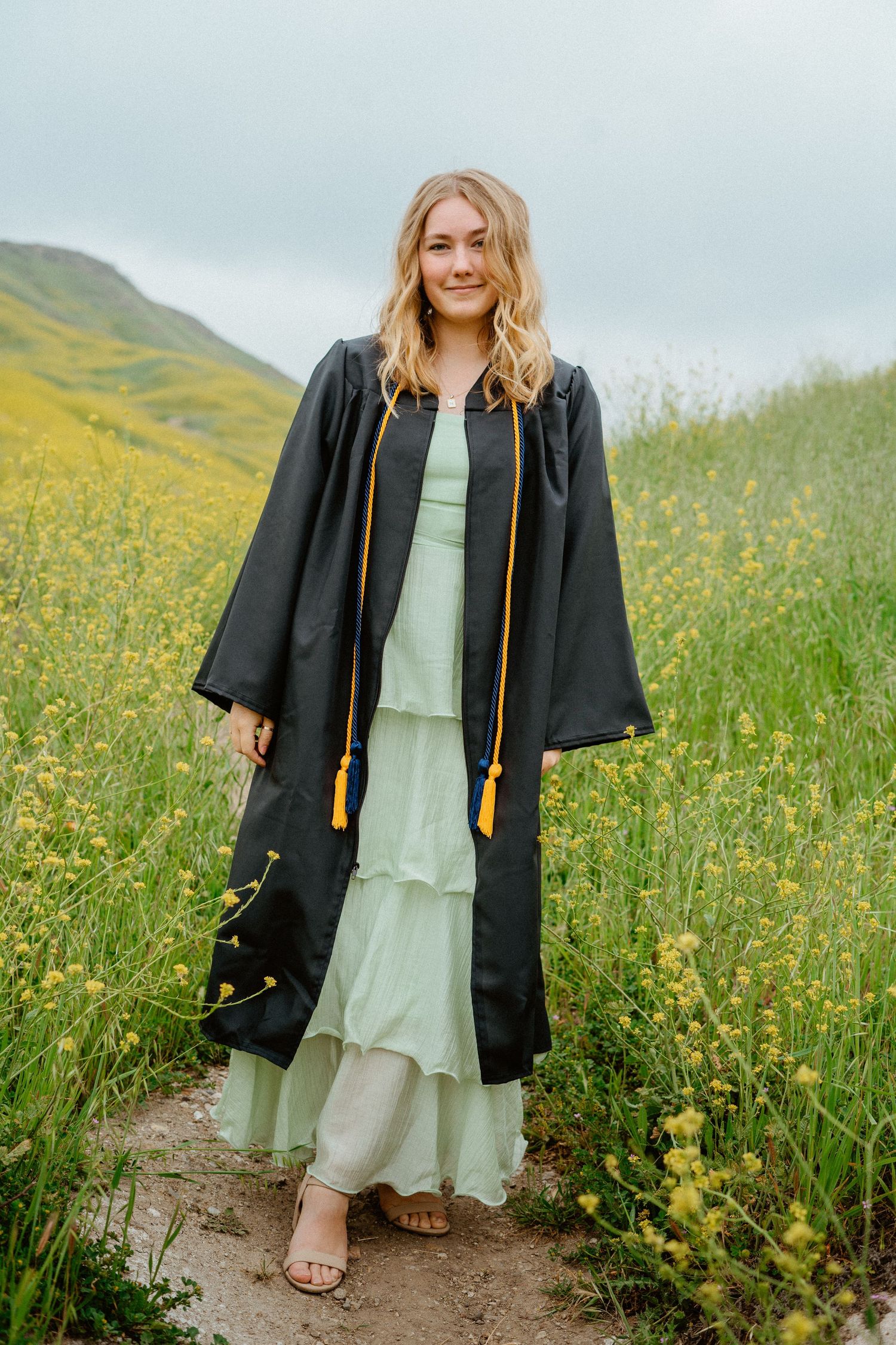 Smiling graduate in black gown and gold tassel over mint dress in flower field.