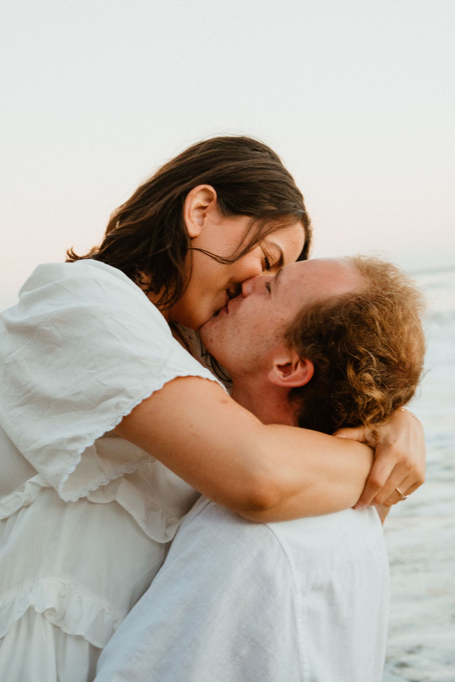 Couple kissing and embracing on beach, woman in white dress hugging man.