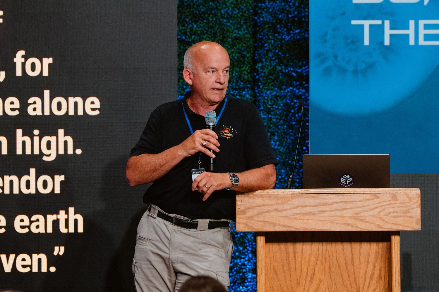Man in black polo shirt speaks at podium with laptop, gesturing during presentation at indoor event.