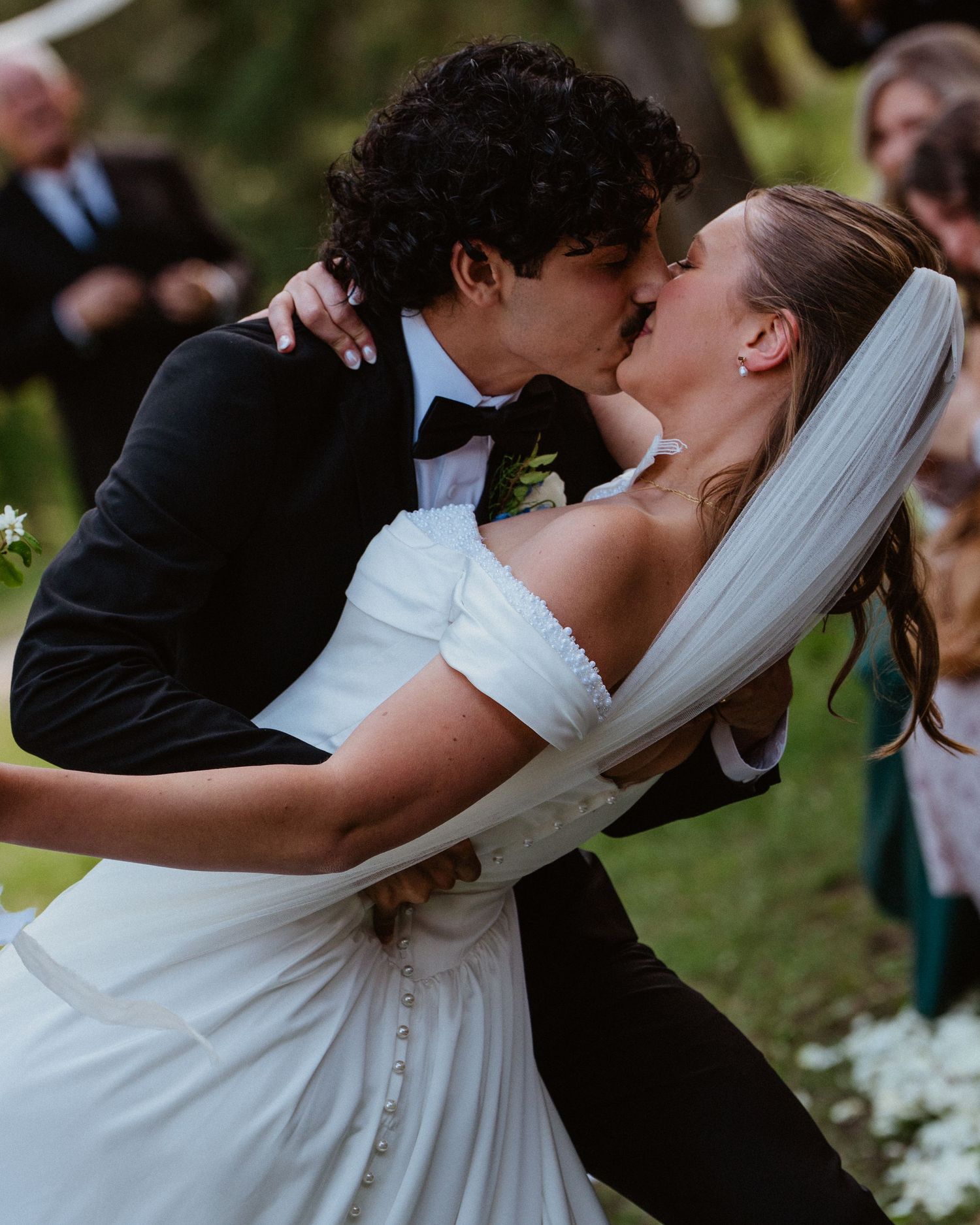 Bride and groom share a passionate first kiss at outdoor wedding ceremony surrounded by guests.