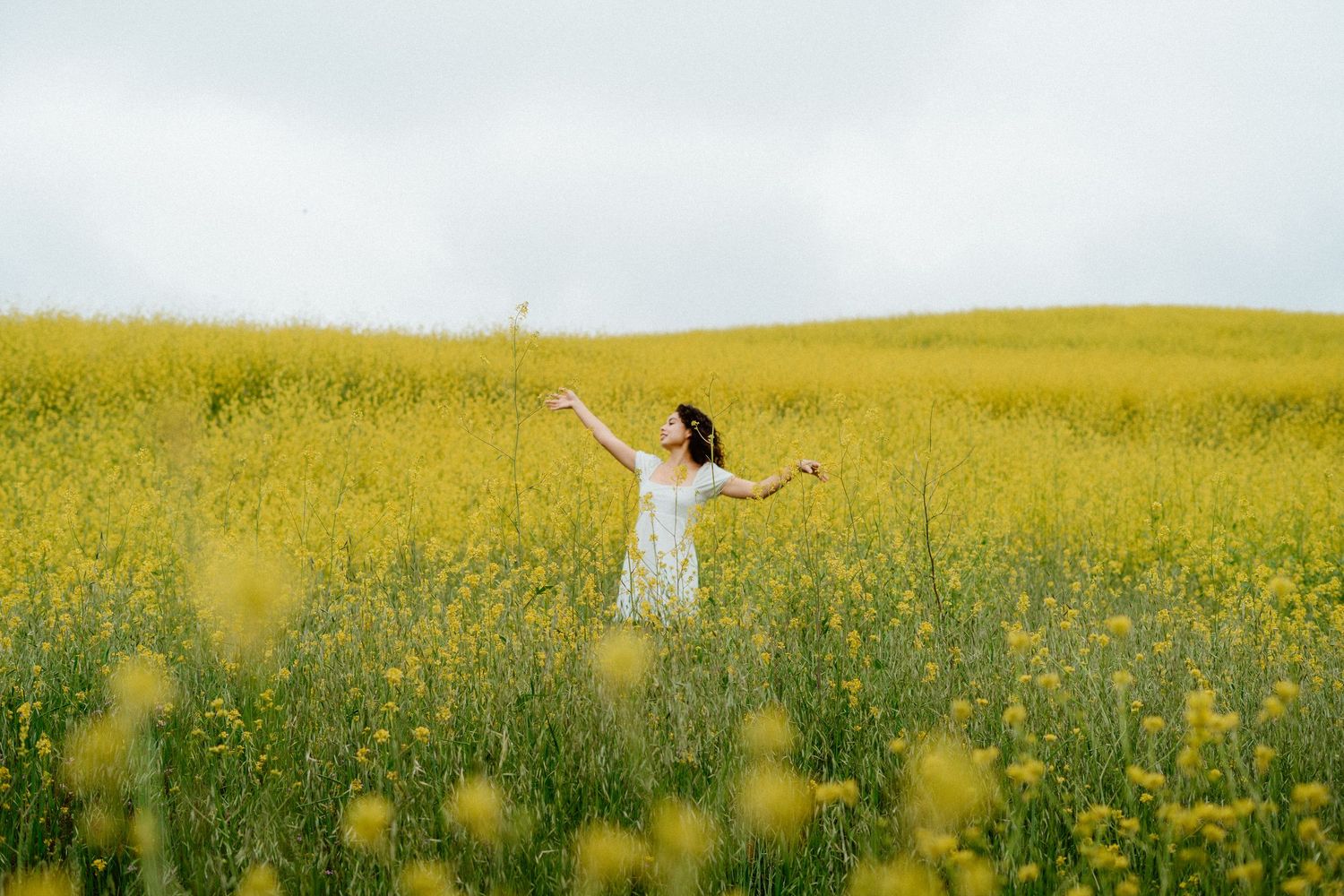 Woman in white dress with arms outstretched joyfully in a vast yellow wildflower field.