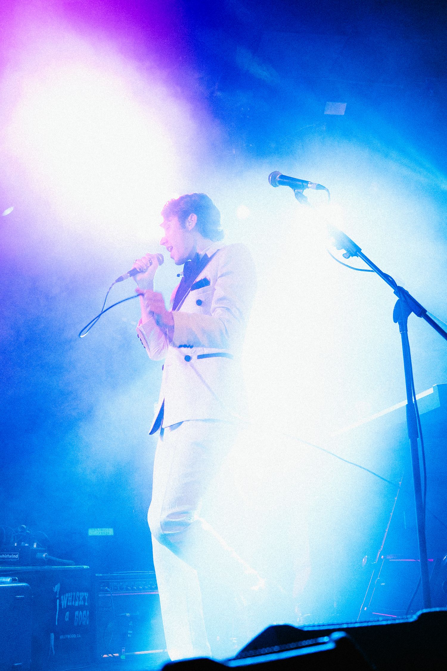 Male performer in white suit singing into microphone on stage with dramatic blue and purple lighting.