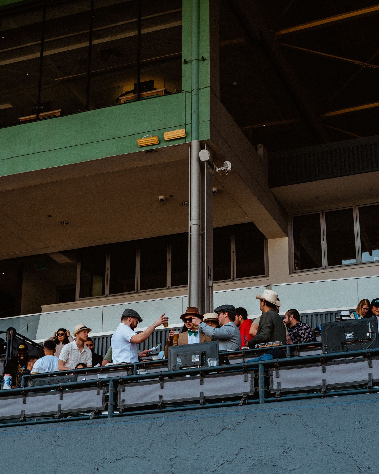 Group of spectators in hats socializing in a stadium grandstand on a sunny day.