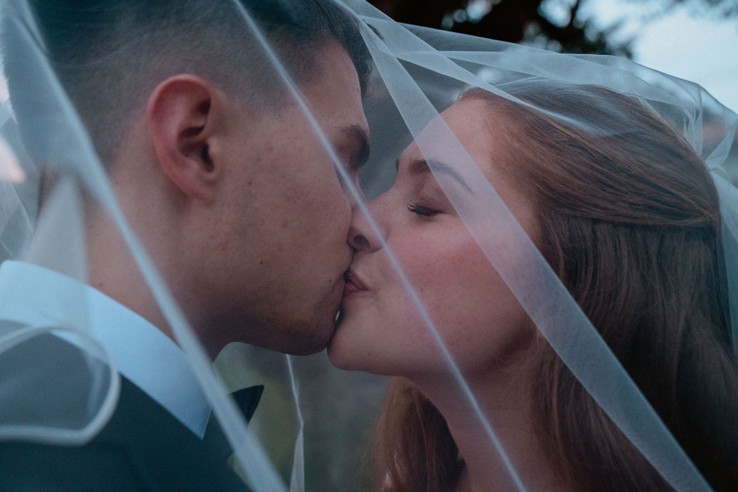 Bride and groom sharing a tender kiss under a flowing white veil on their wedding day.