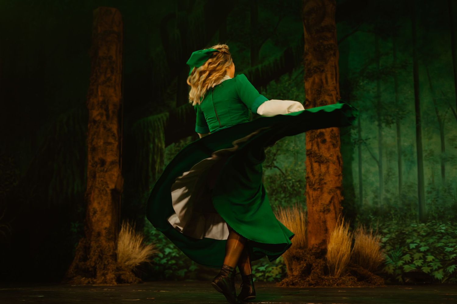 A dancer in a green costume twirls gracefully against a forest-themed stage backdrop.