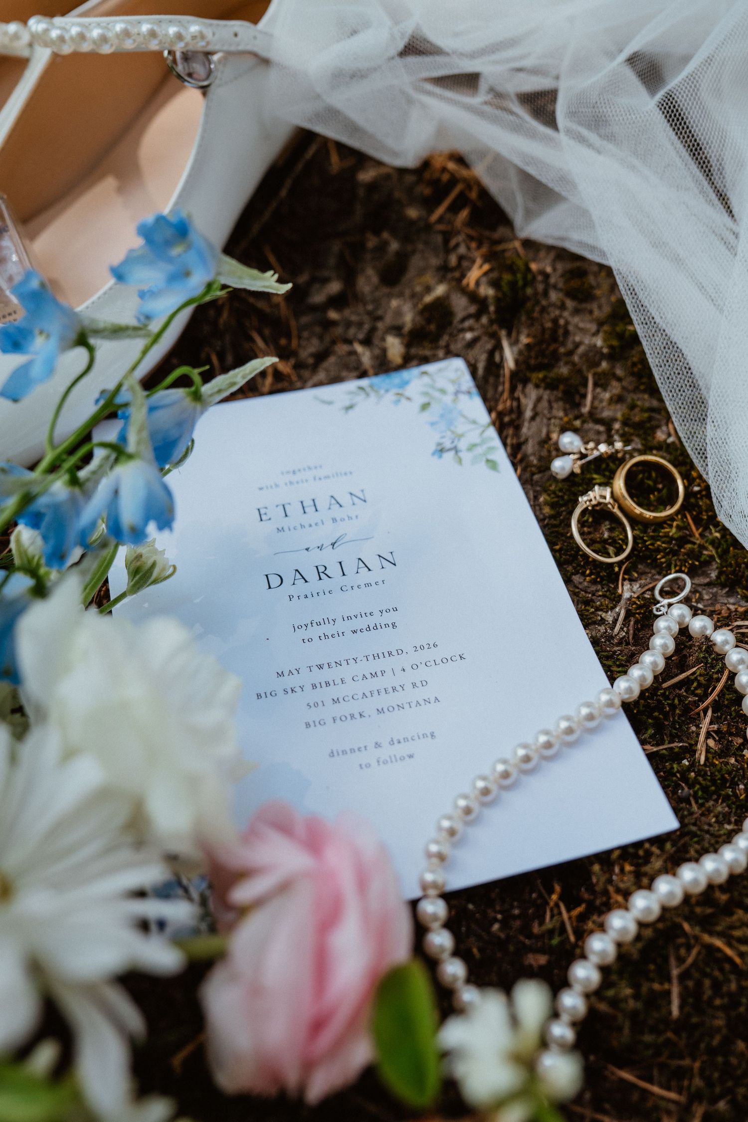 Close-up wedding flat lay featuring floral invitation for Ethan and Darian, pearl necklace, and gold rings.
