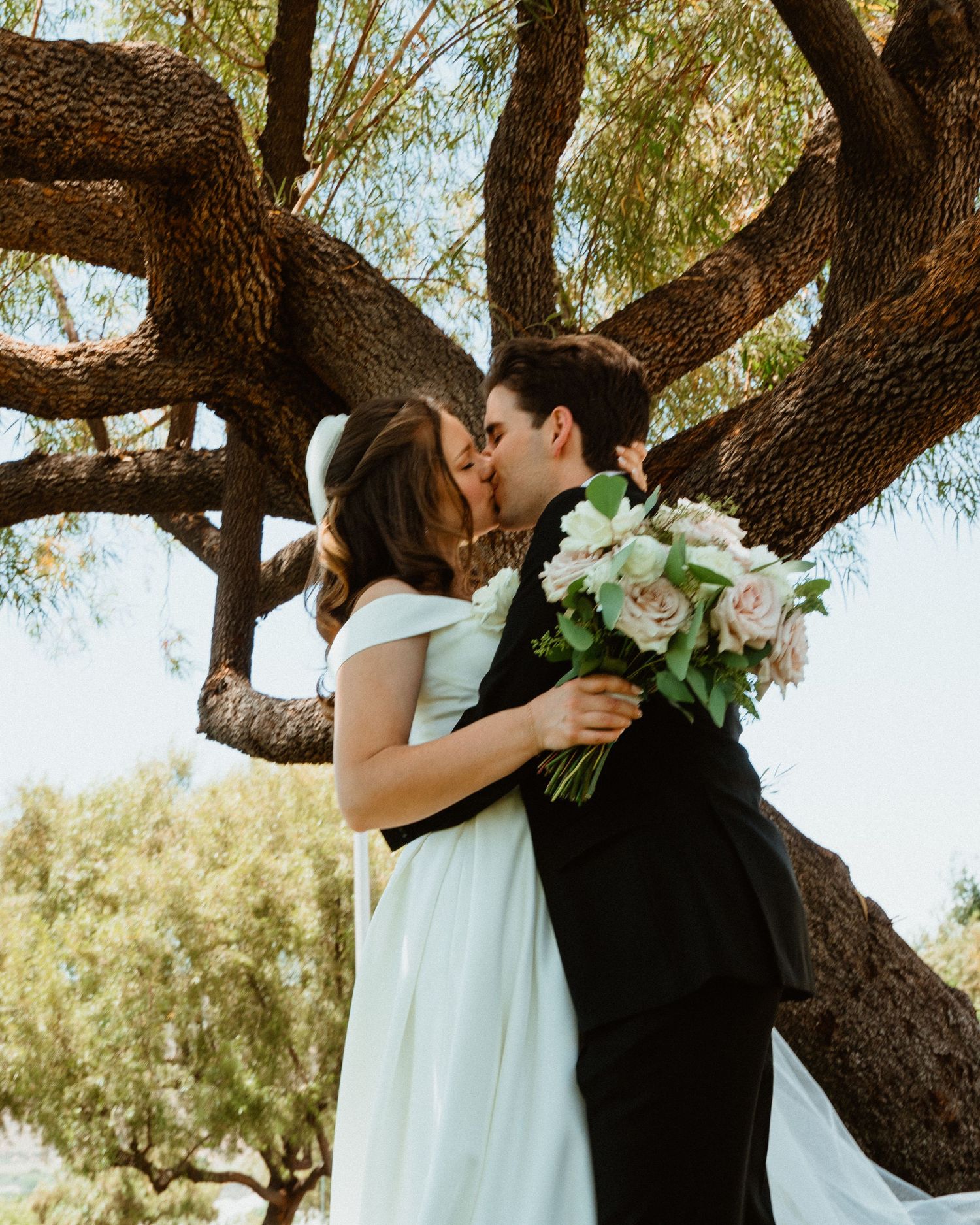 Newlyweds share a romantic kiss under a sprawling tree, bride holding a blush bouquet in a white gown.