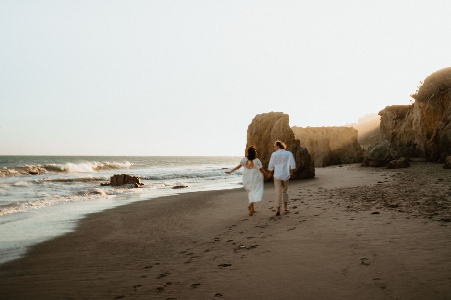 Couple holding hands walking on a sandy beach at sunset near large rocky cliffs with ocean waves.