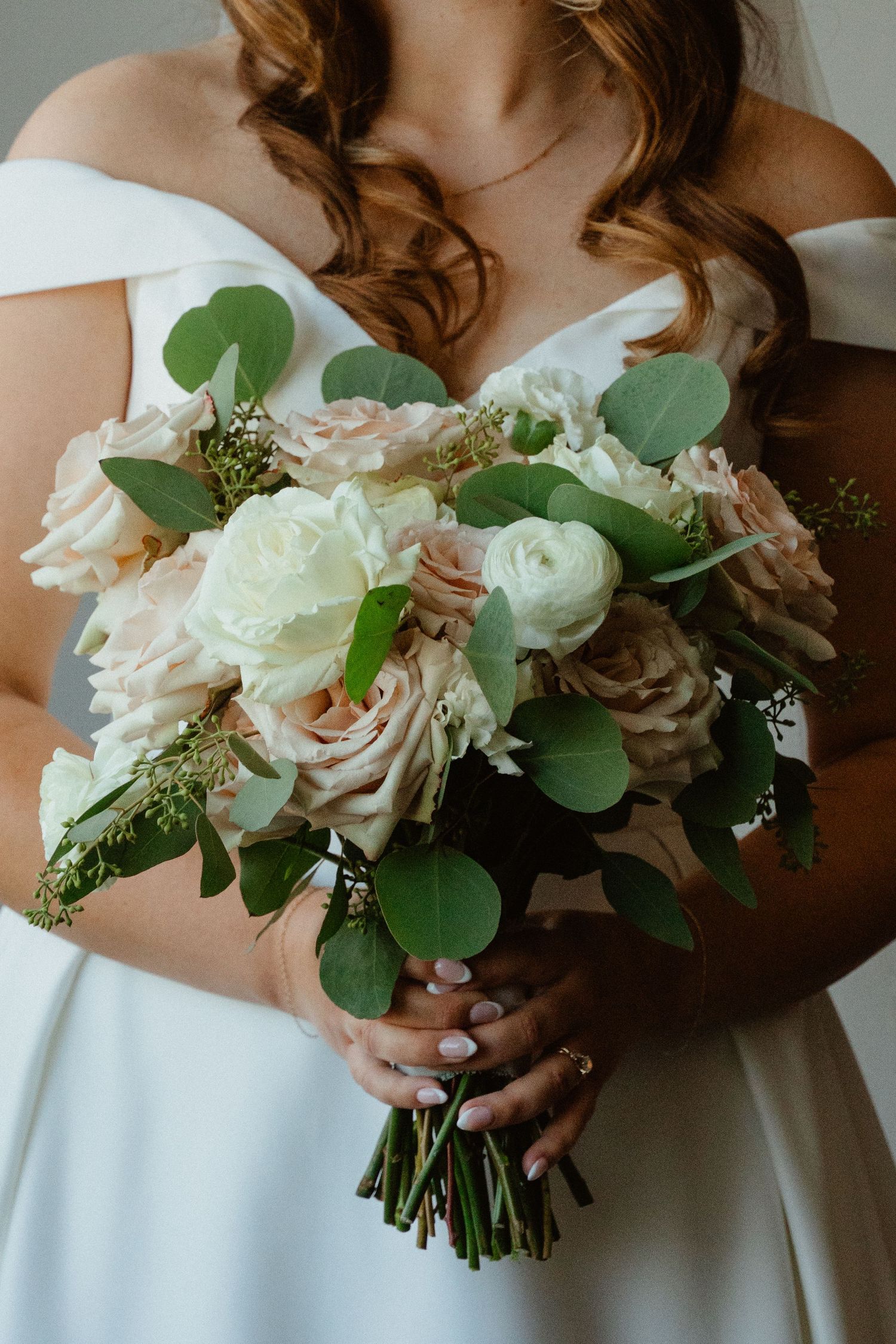 Bride holding a wedding bouquet of blush and white roses with eucalyptus greenery in an off-shoulder gown.