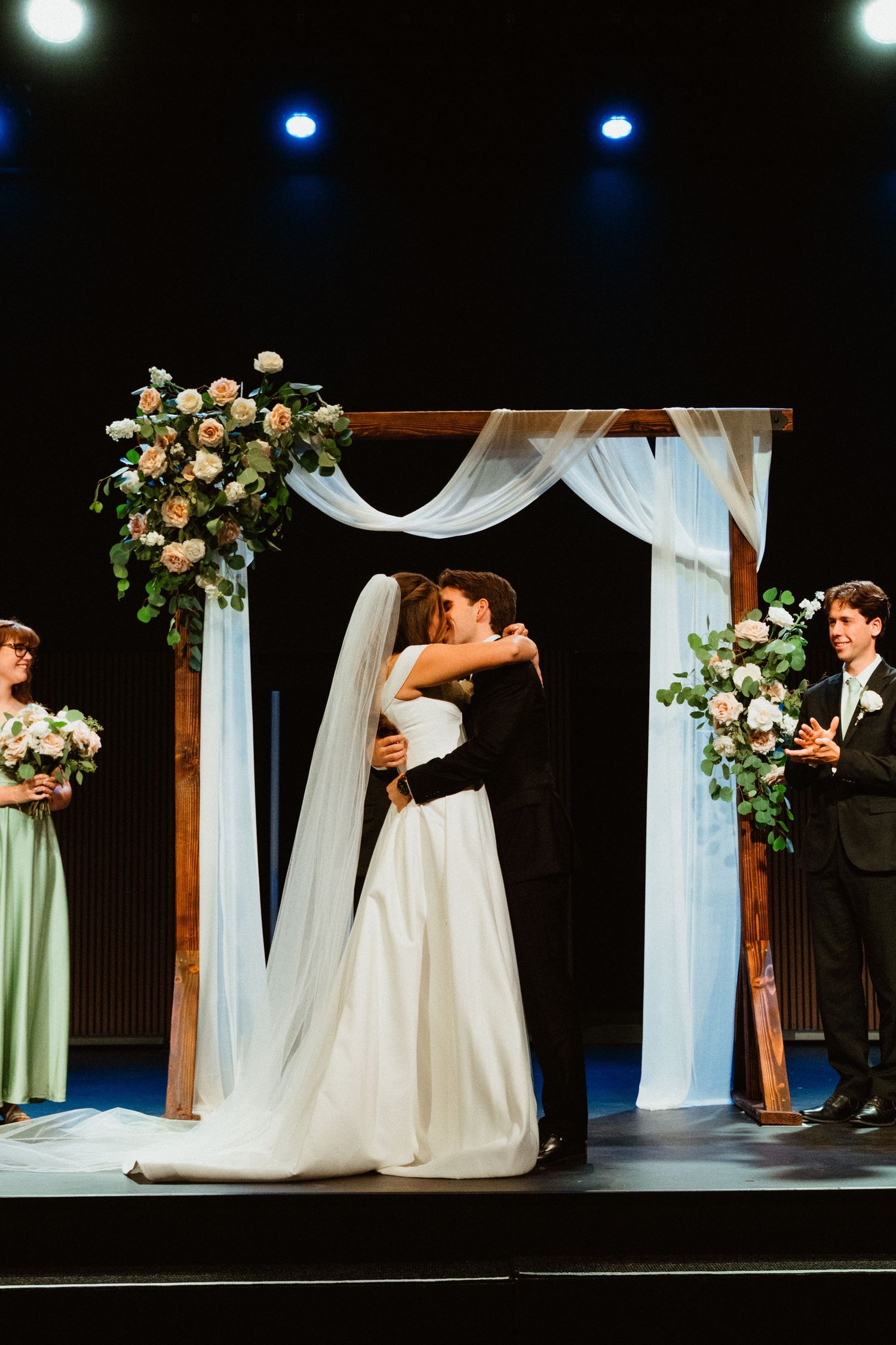 Bride and groom share first kiss under rustic wooden arch with white draping and floral arrangements at wedding ceremony.