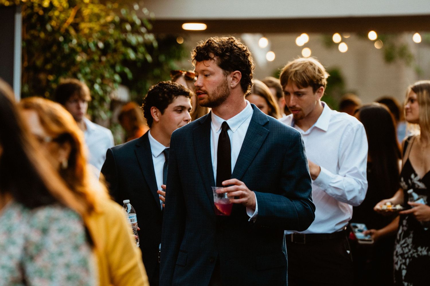 Men in suits socializing at an elegant outdoor evening party, holding drinks amid a crowd of guests.