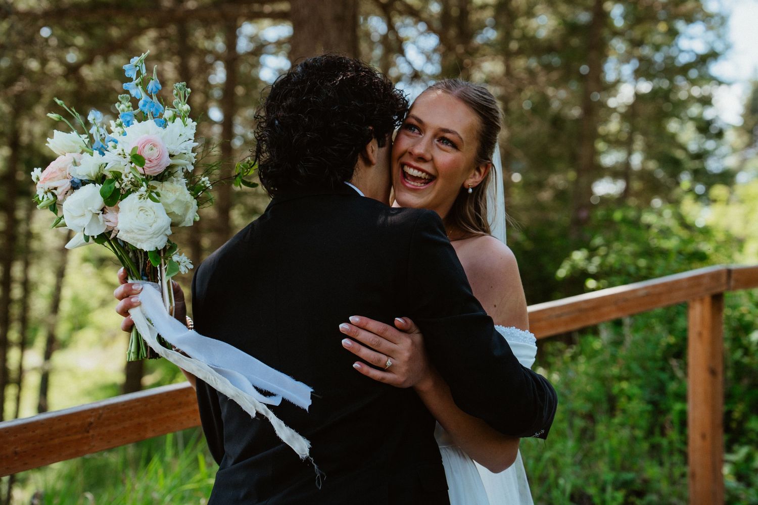 Bride in white dress laughing while embracing groom in black suit, holding blue and pink floral bouquet outdoors.