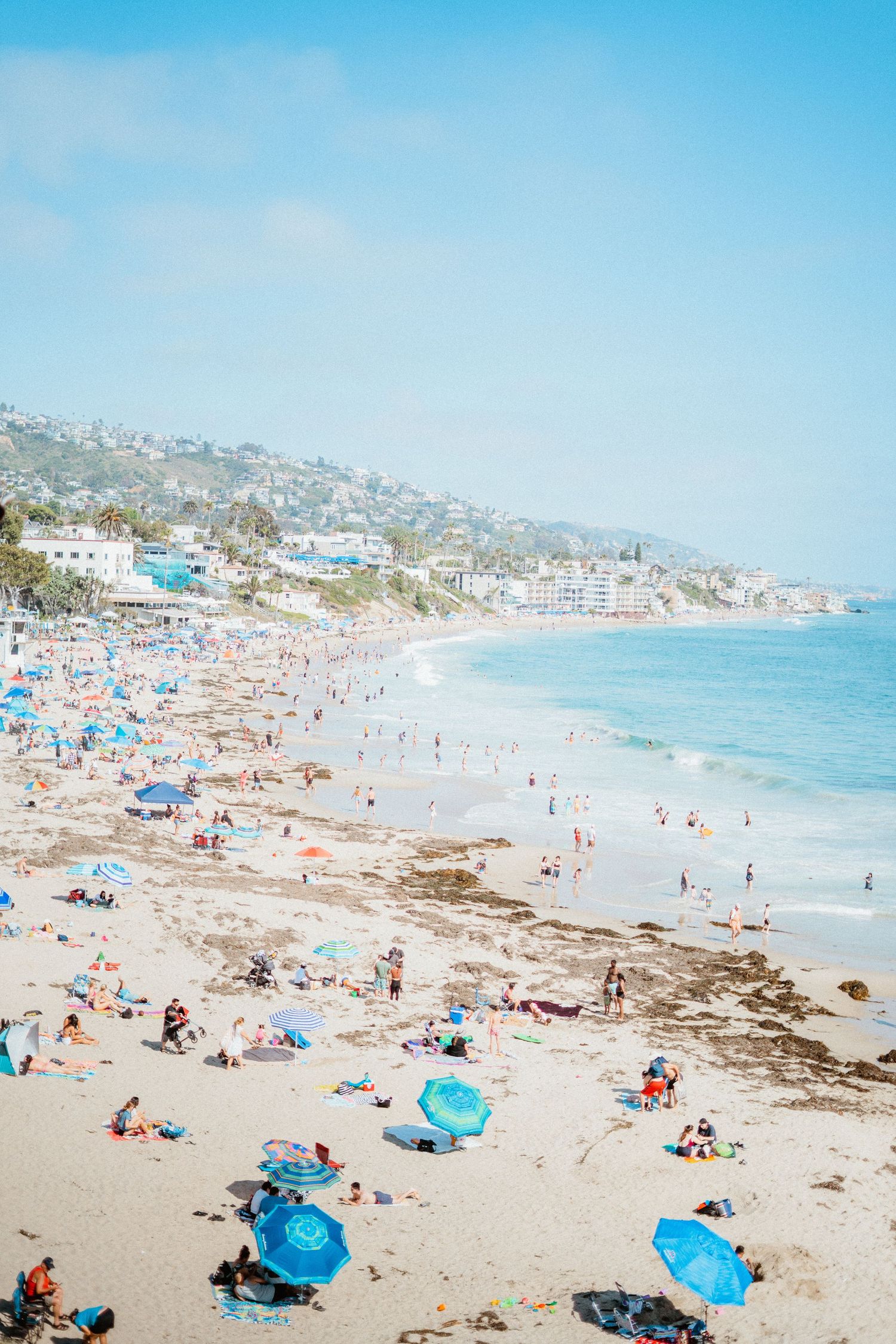 Crowded sunny beach with blue umbrellas and coastal town in background on clear day.