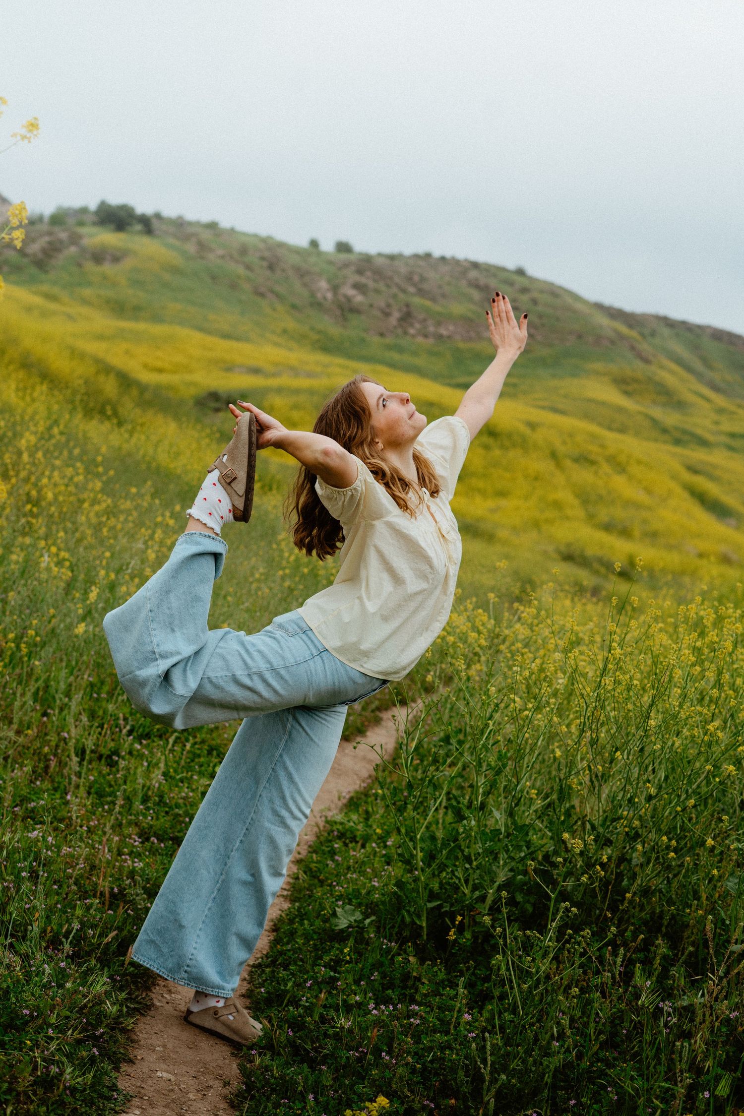 Woman in white top and blue flared jeans doing a yoga dancer pose in a green meadow.