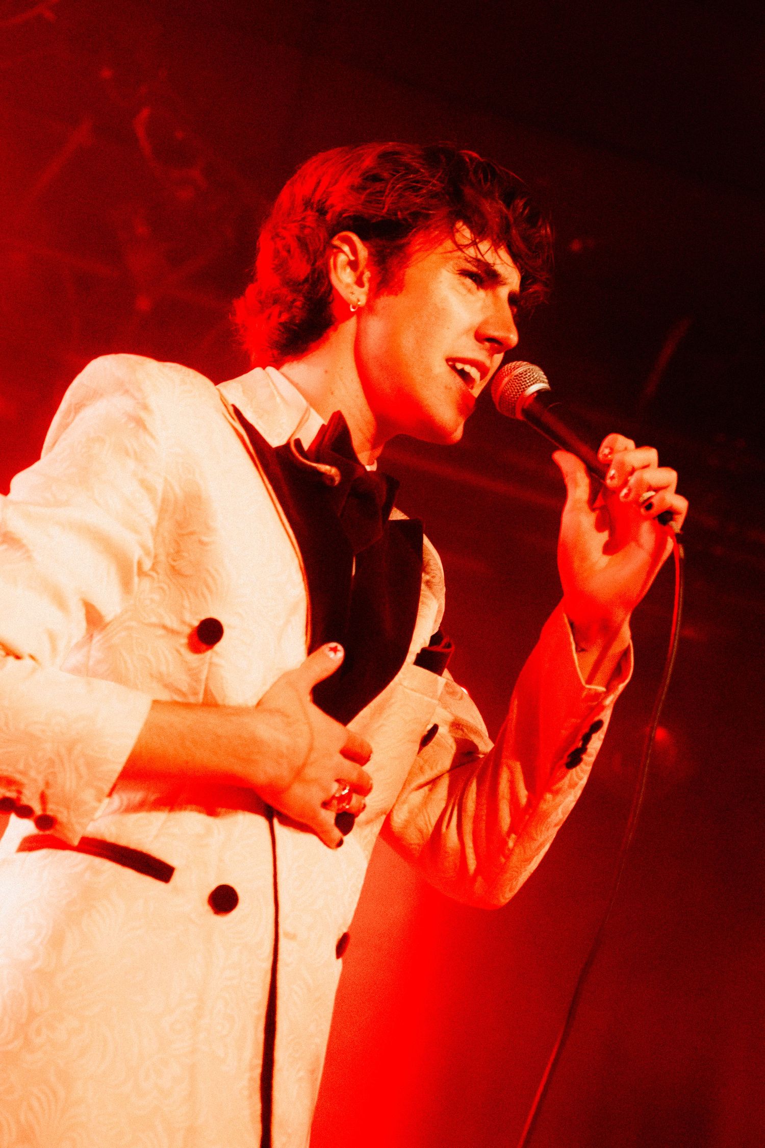 Young male singer in white double-breasted suit performing with microphone under red stage lights.