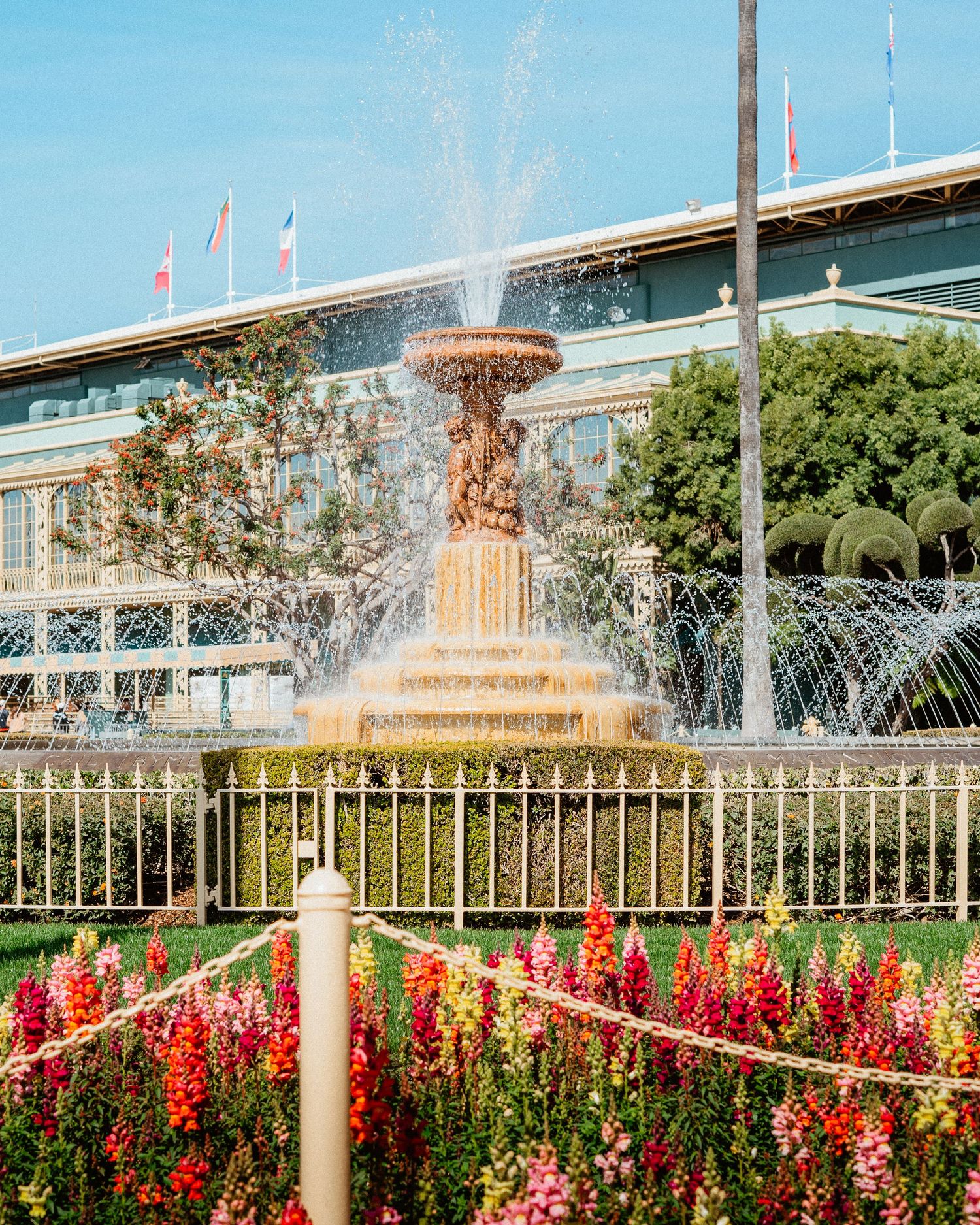 Ornate fountain surrounded by colorful flowers and lush greenery at a sunny racetrack.