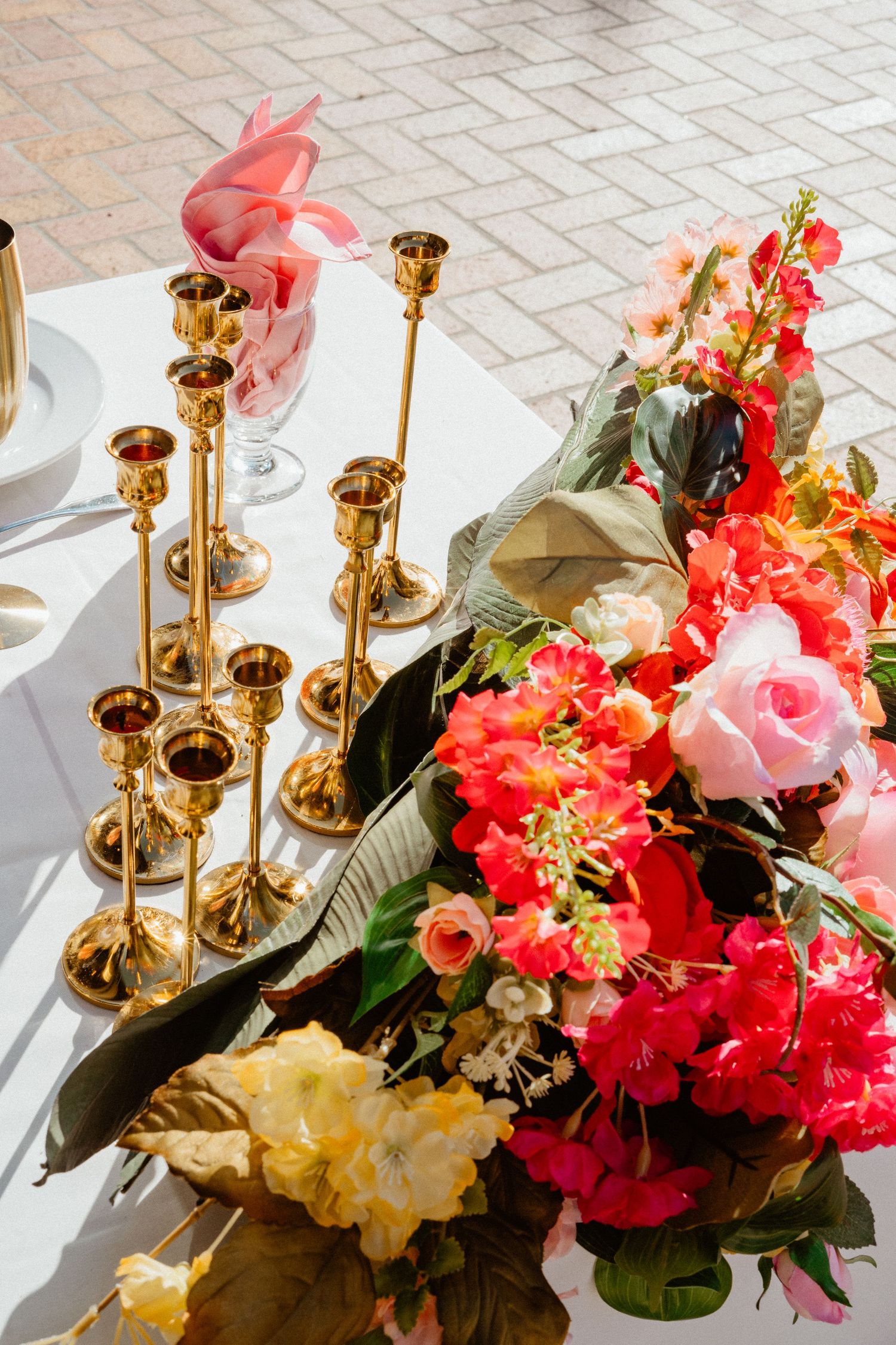 Gold brass candlestick holders arranged on white wedding table beside vibrant red, pink, and yellow floral centerpiece.