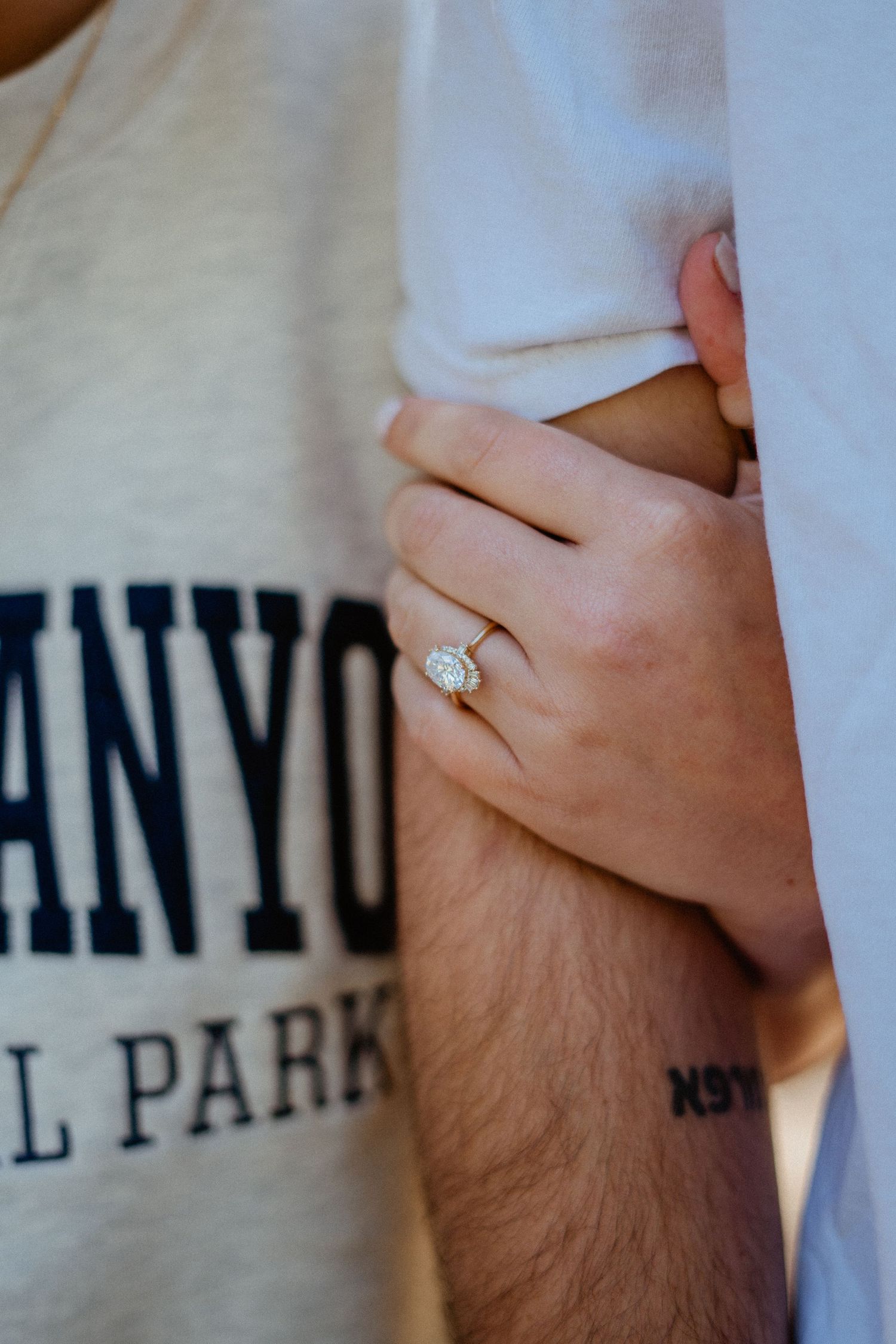Close-up of oval diamond engagement ring on womans hand holding mans arm outdoors.