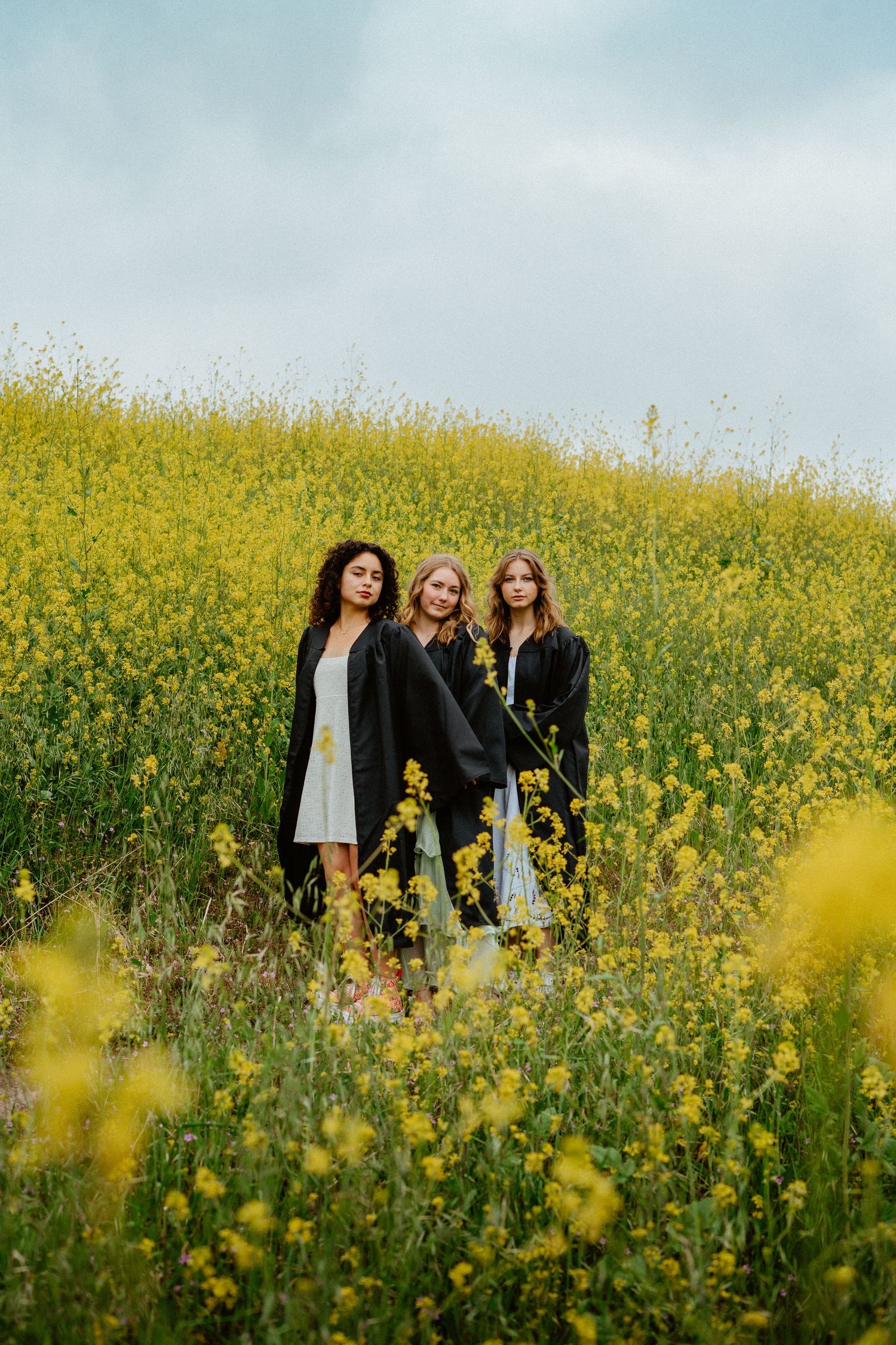 Three young women in black blazers posing together in a stunning yellow wildflower meadow under cloudy skies.