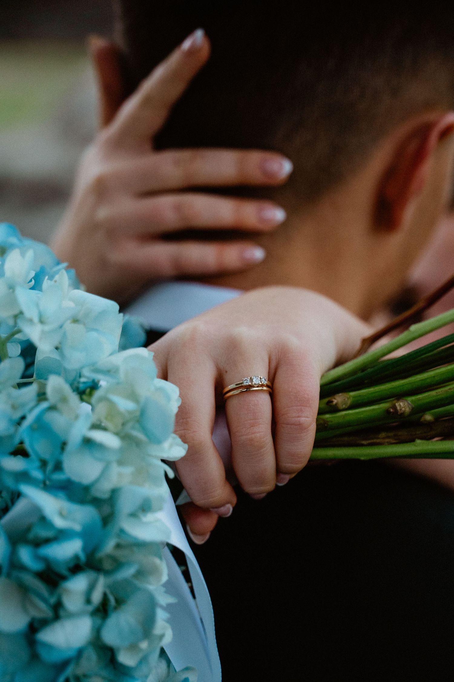 Close-up of bride's wedding ring and hand holding blue hydrangea bouquet embracing the groom.