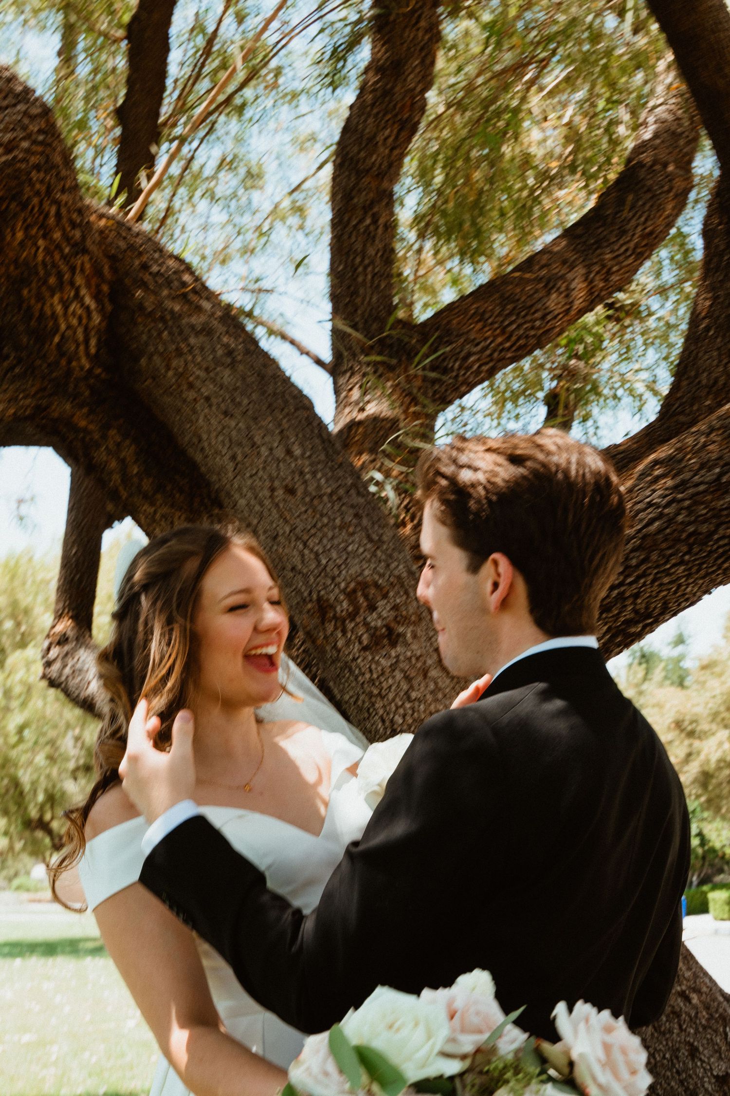 Joyful bride laughing as groom holds her face near a large tree outdoors on their wedding day.