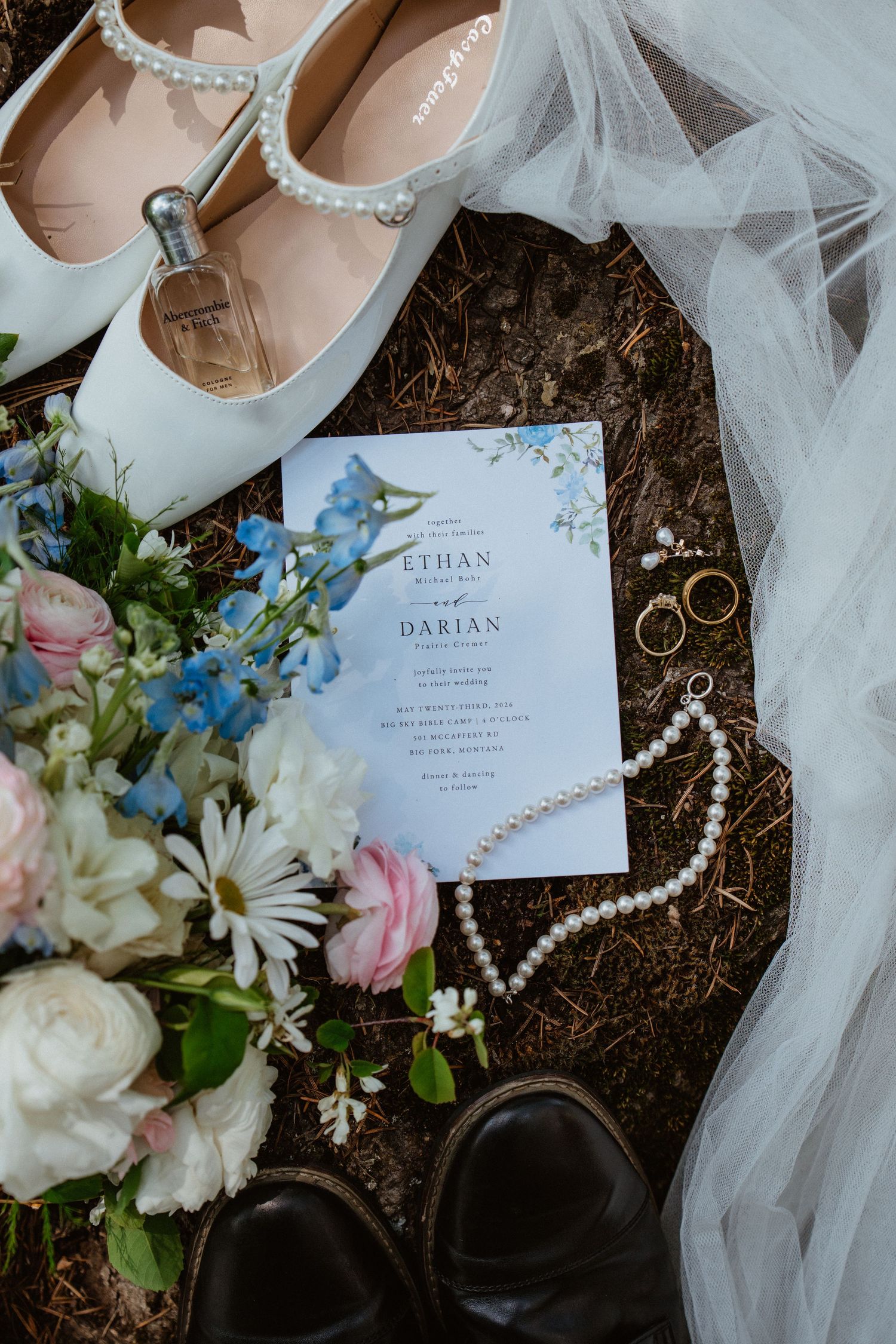 Wedding flat lay with invitation, pearl necklace, rings, bridal heels, bouquet, and veil on rustic ground.