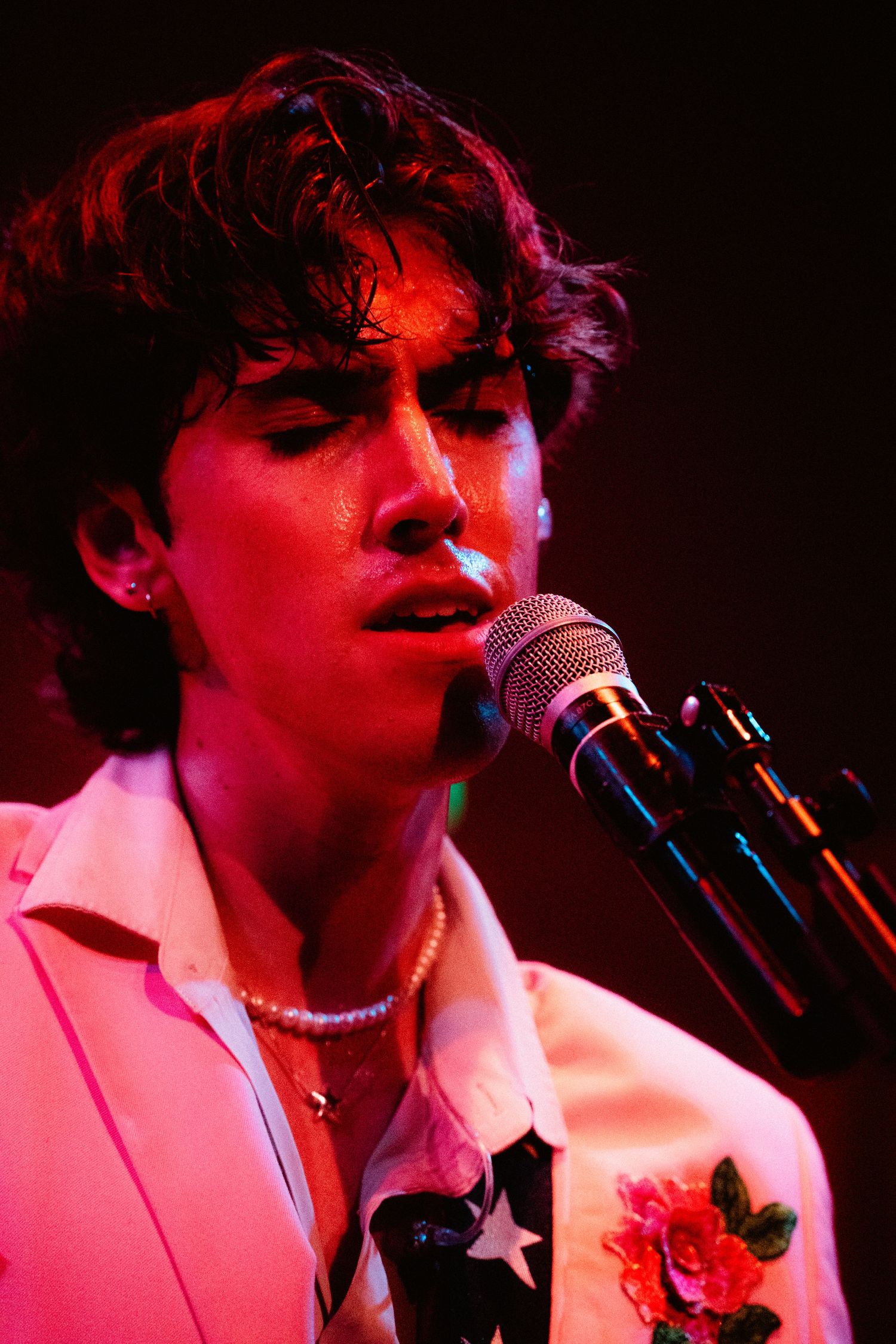 Close-up of young male singer performing at microphone under warm red stage lighting.
