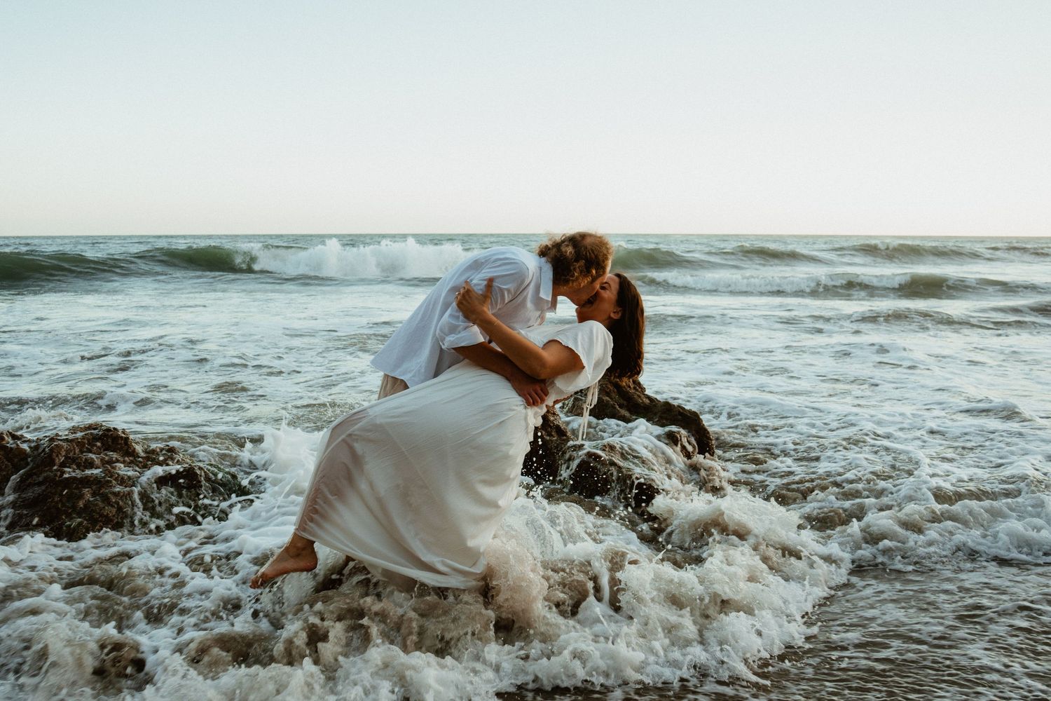 Couple in wedding attire kissing romantically in ocean waves on a rocky beach at sunset.