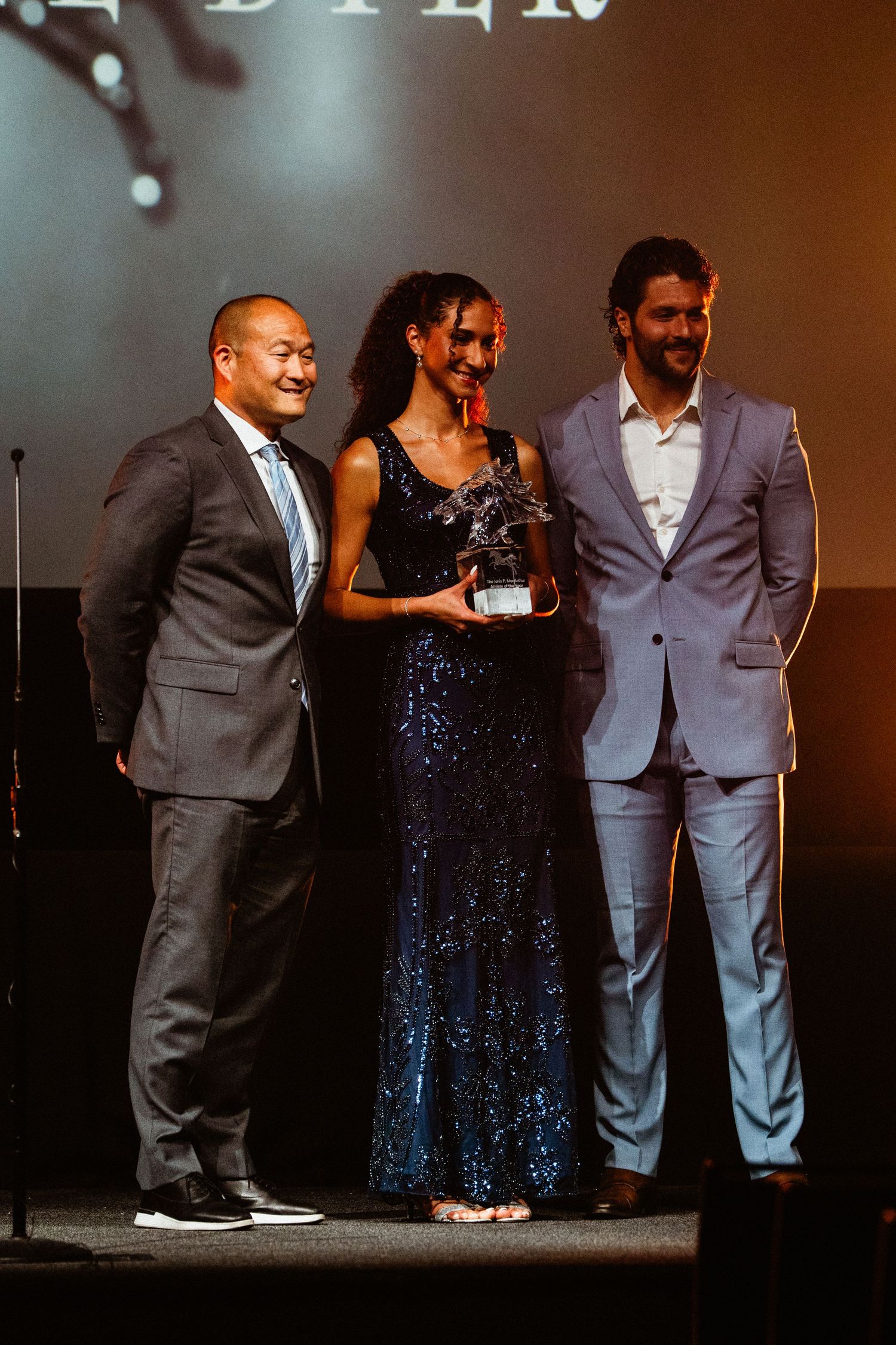 Three people posing on stage at awards ceremony, woman in sequin gown holding trophy.