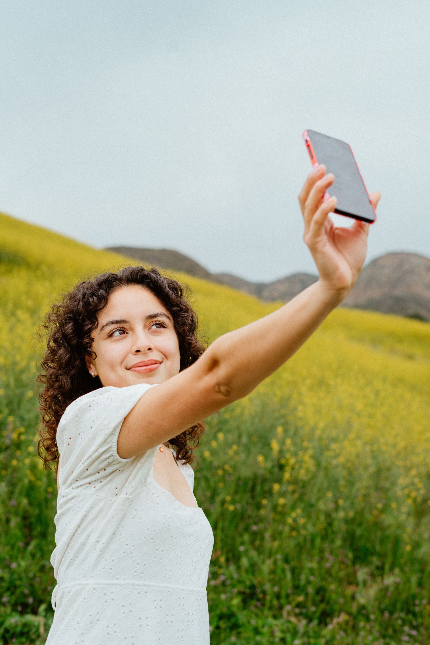 Young woman with curly hair taking a selfie in a vibrant yellow flower field with hills in the background.