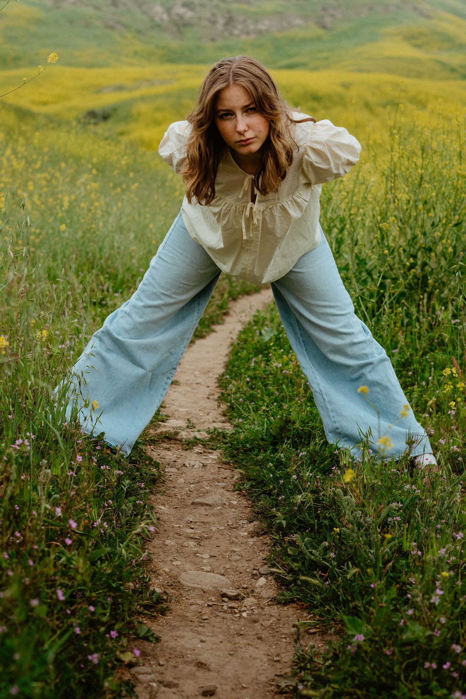 Young woman in white blouse and wide-leg jeans posing on a wildflower meadow trail.