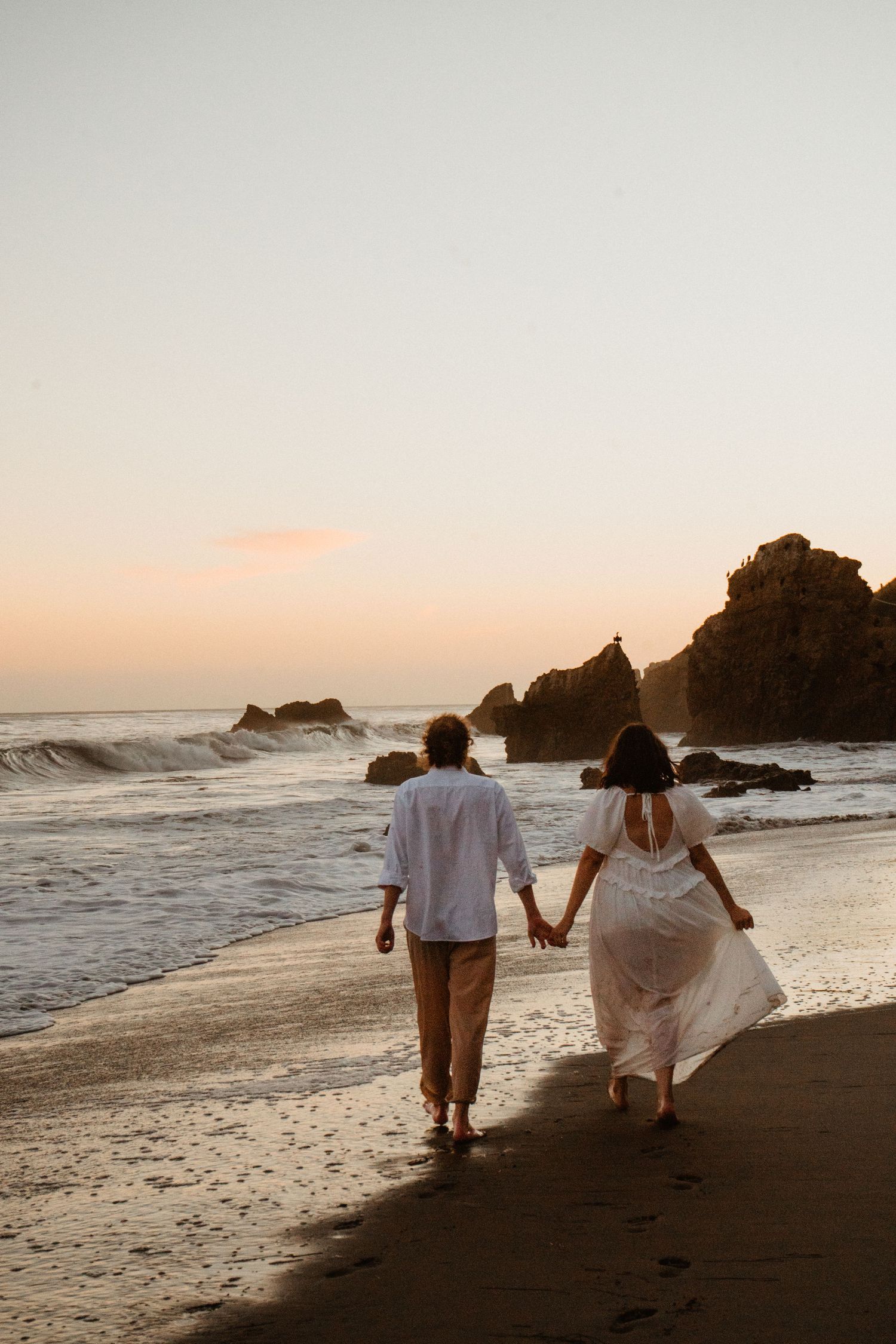 Couple holding hands walking along a sandy beach at sunset with ocean waves and rocky cliffs in the background.