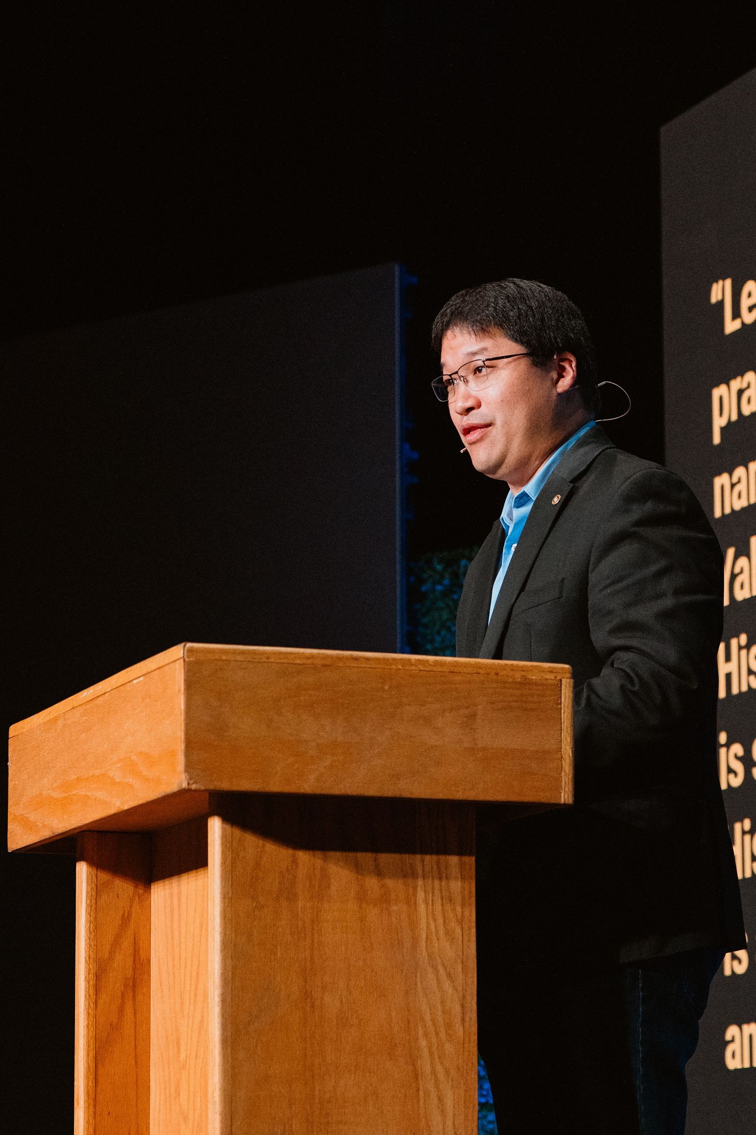 Man in suit speaking at wooden podium during formal presentation with text display in background.