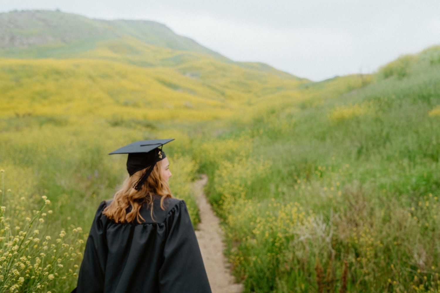 Graduate in black cap and gown walks a wildflower trail with lush yellow hills in the background.