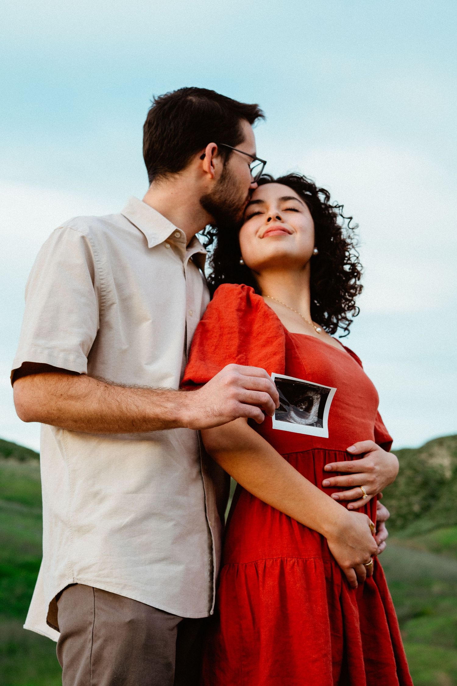 Man kissing woman's head while embracing outdoors, both holding ultrasound photo on scenic hillside.