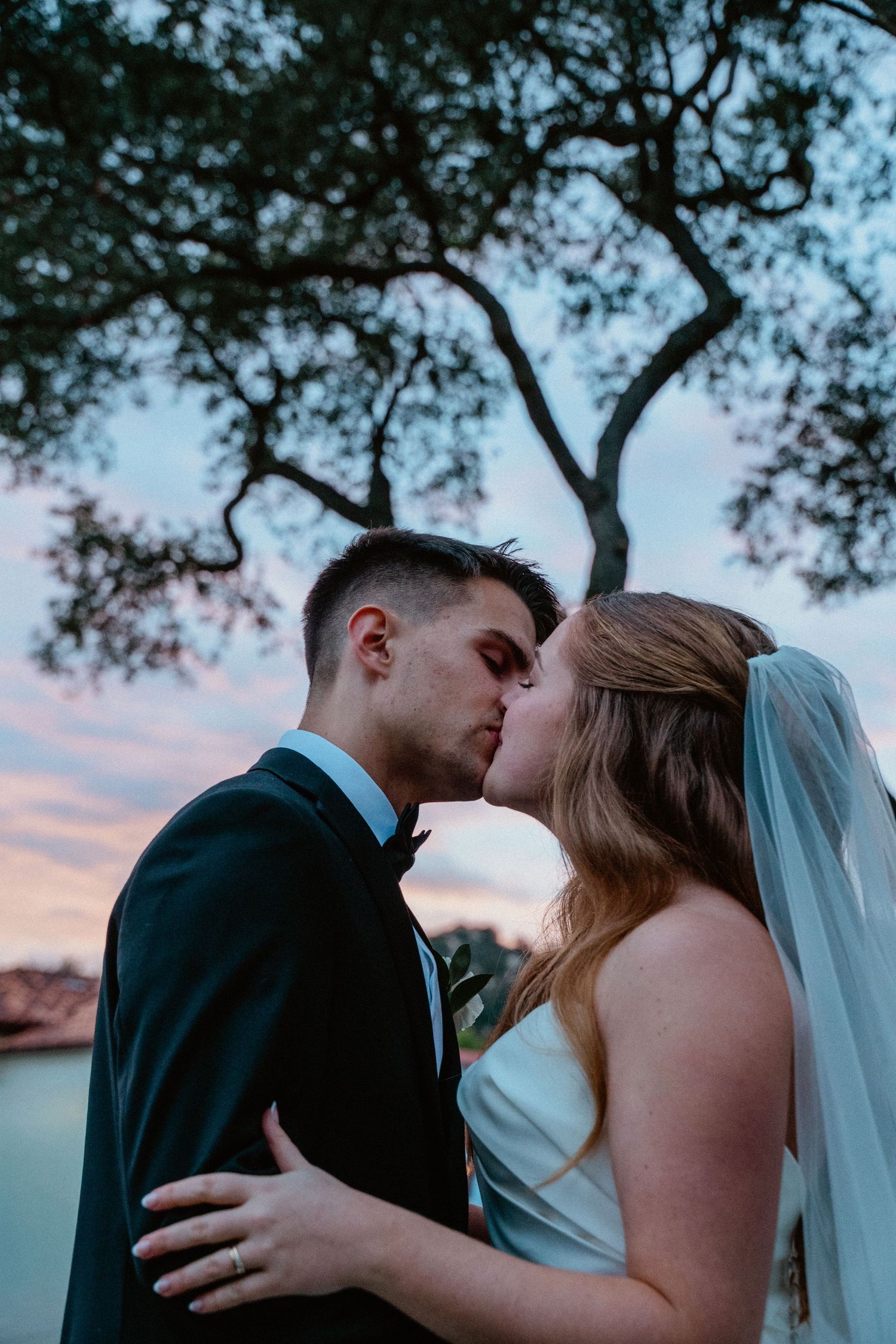 Bride and groom sharing a romantic kiss under a tree at sunset on their wedding day.