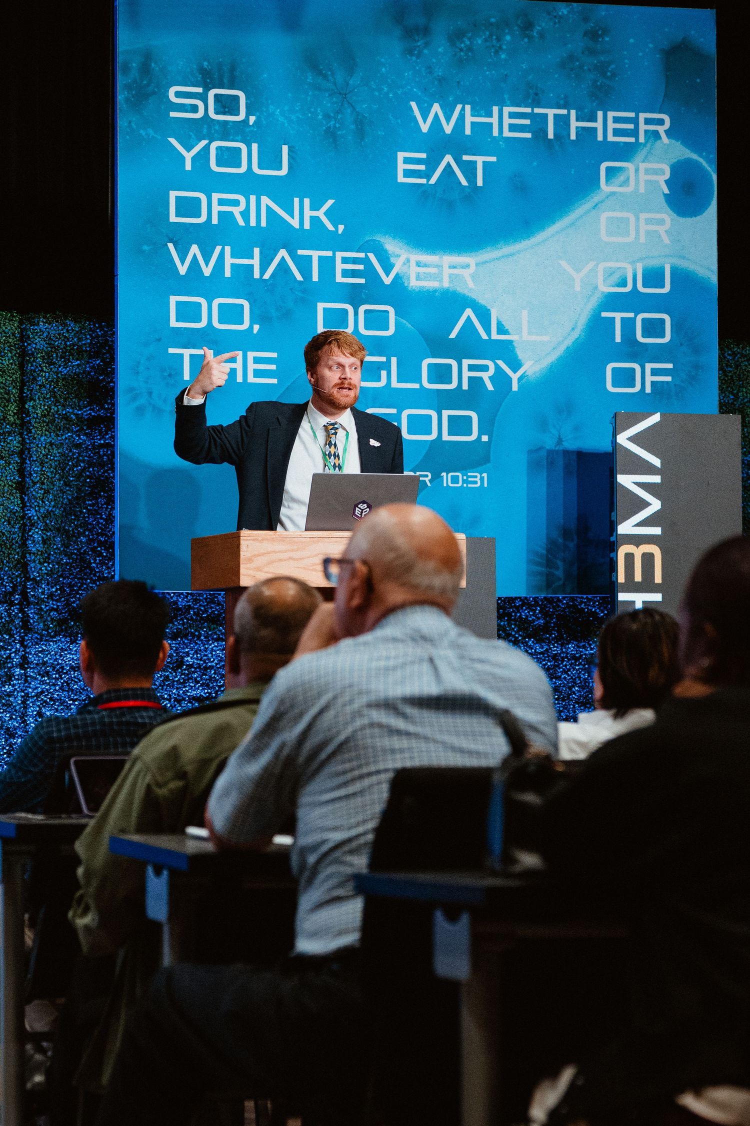 A speaker at a podium gestures while presenting a Bible verse about glorifying God on a blue projection screen.