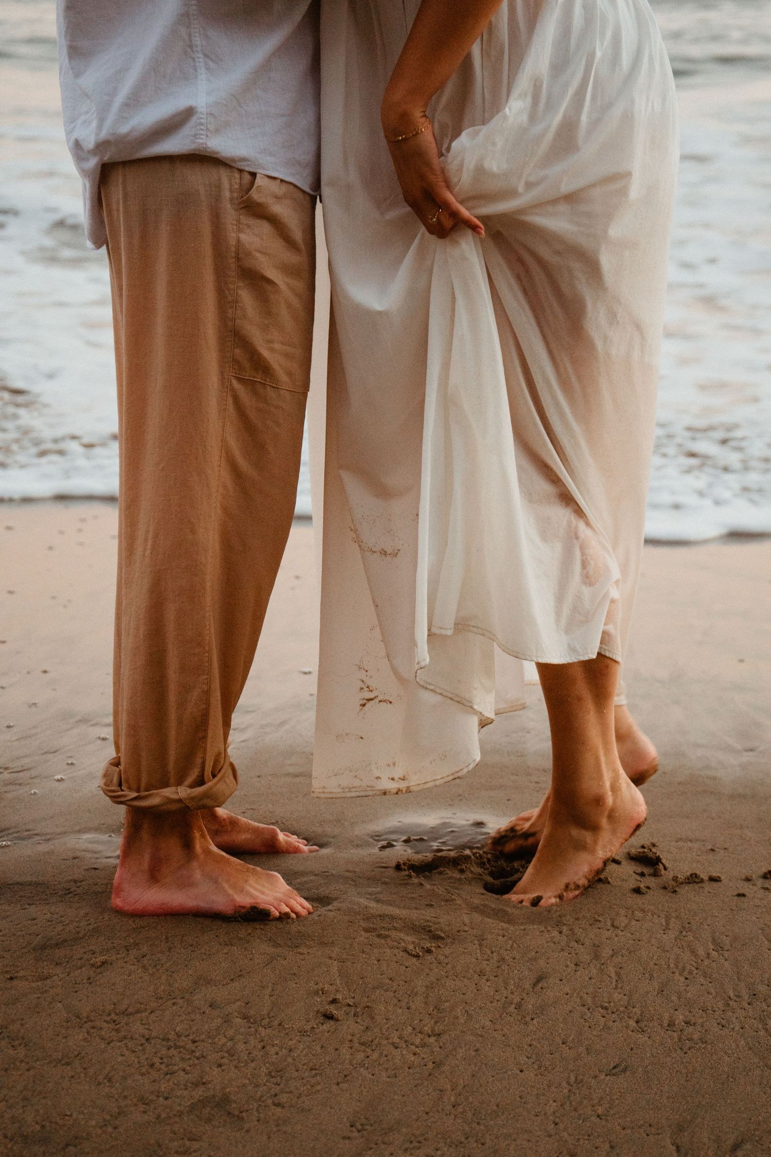 Close-up of barefoot couple standing on wet sand, woman in white dress, man in beige rolled-up pants.