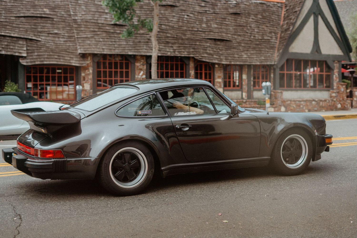 Vintage dark gray Porsche 911 Turbo parked on street near Tudor-style building.