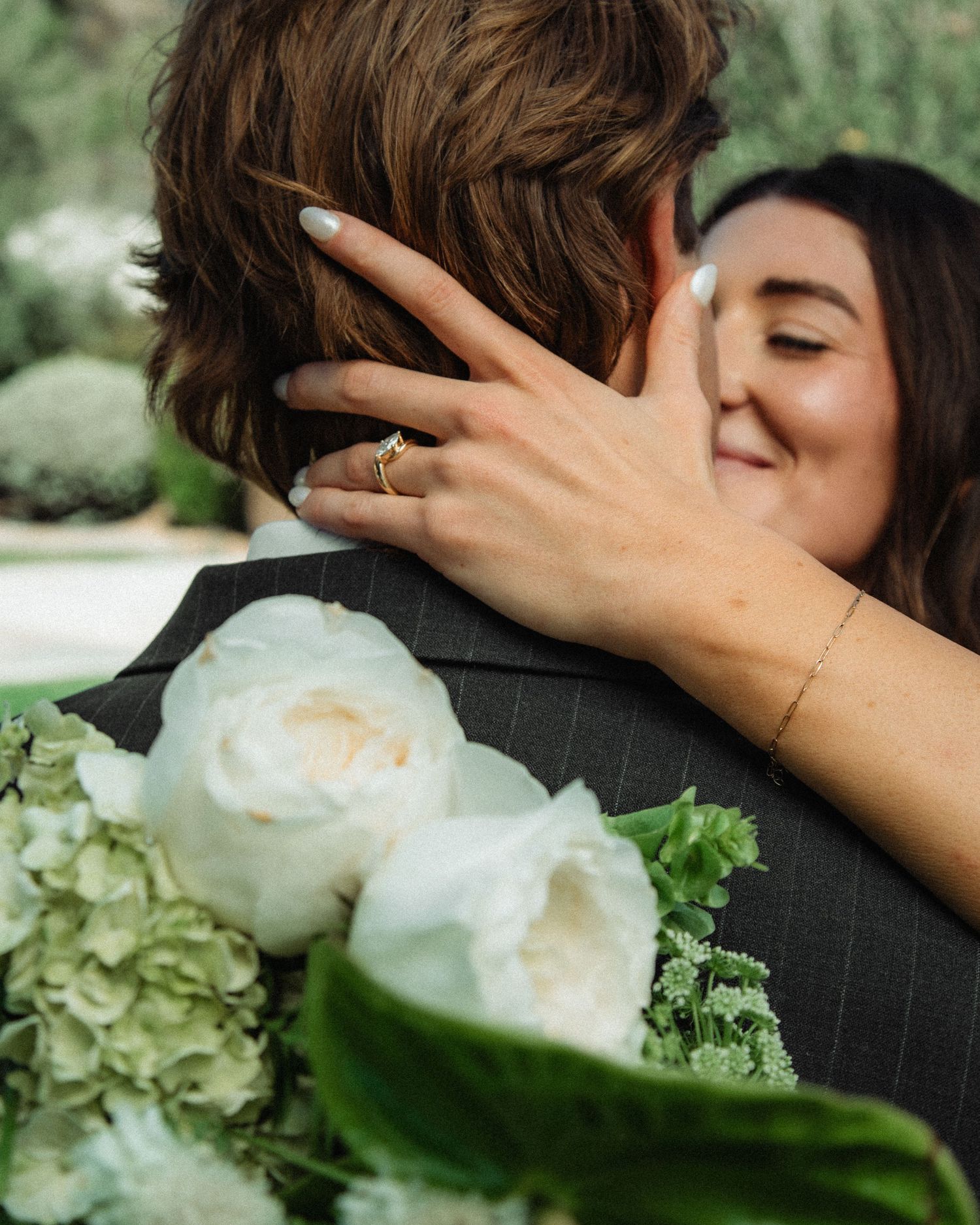 A bride with mint nails and a gold ring embracing her groom while holding white wedding flowers.