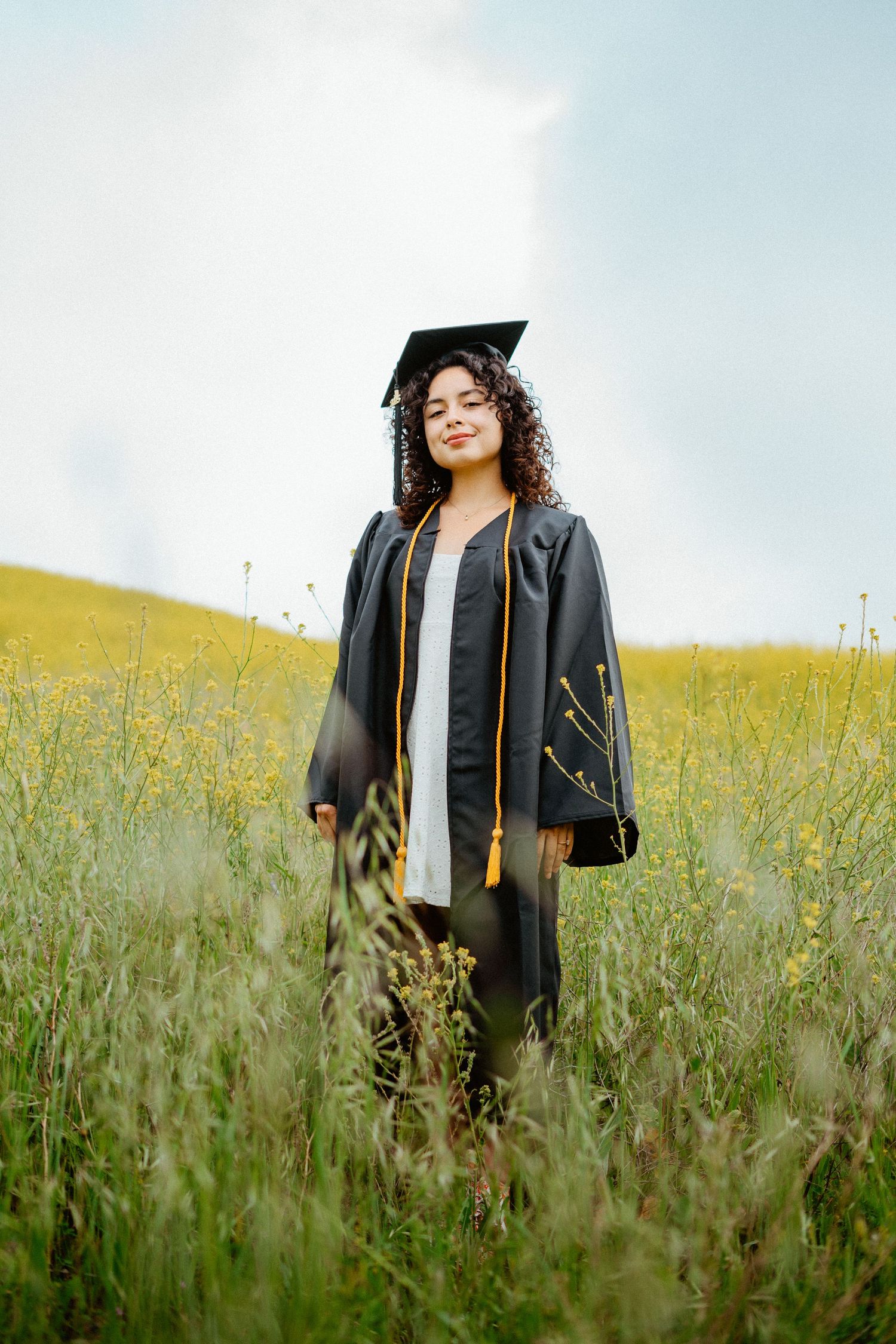 Graduate in black cap and gown standing in a lush yellow wildflower meadow.