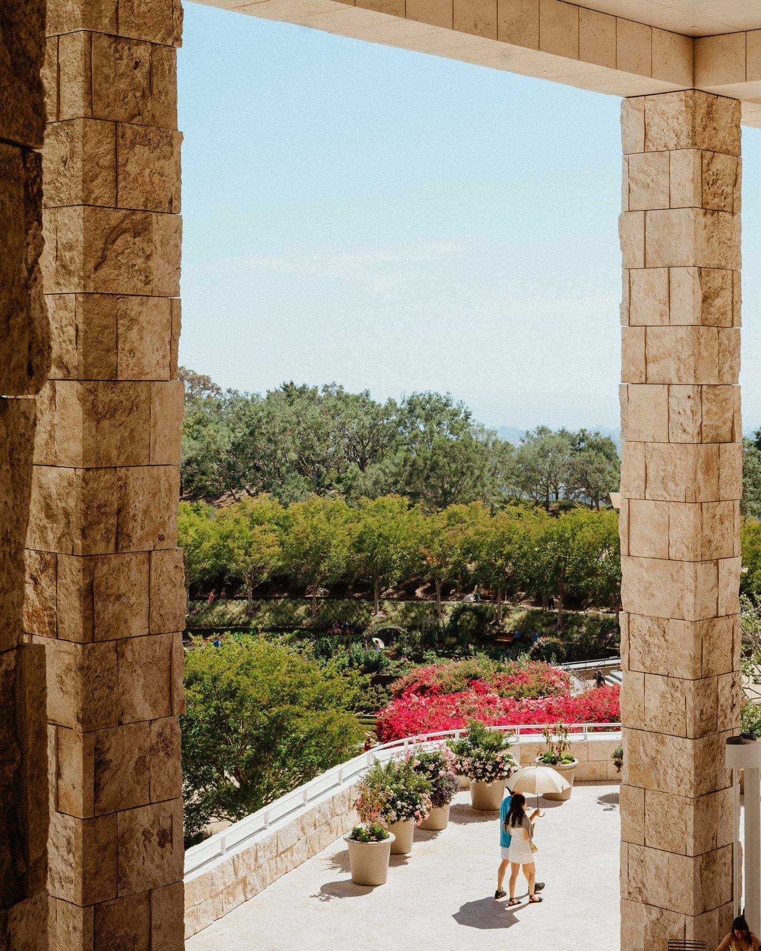 Two visitors walk through stone colonnade archway overlooking lush garden terrace with colorful bougainvillea and ocean view.