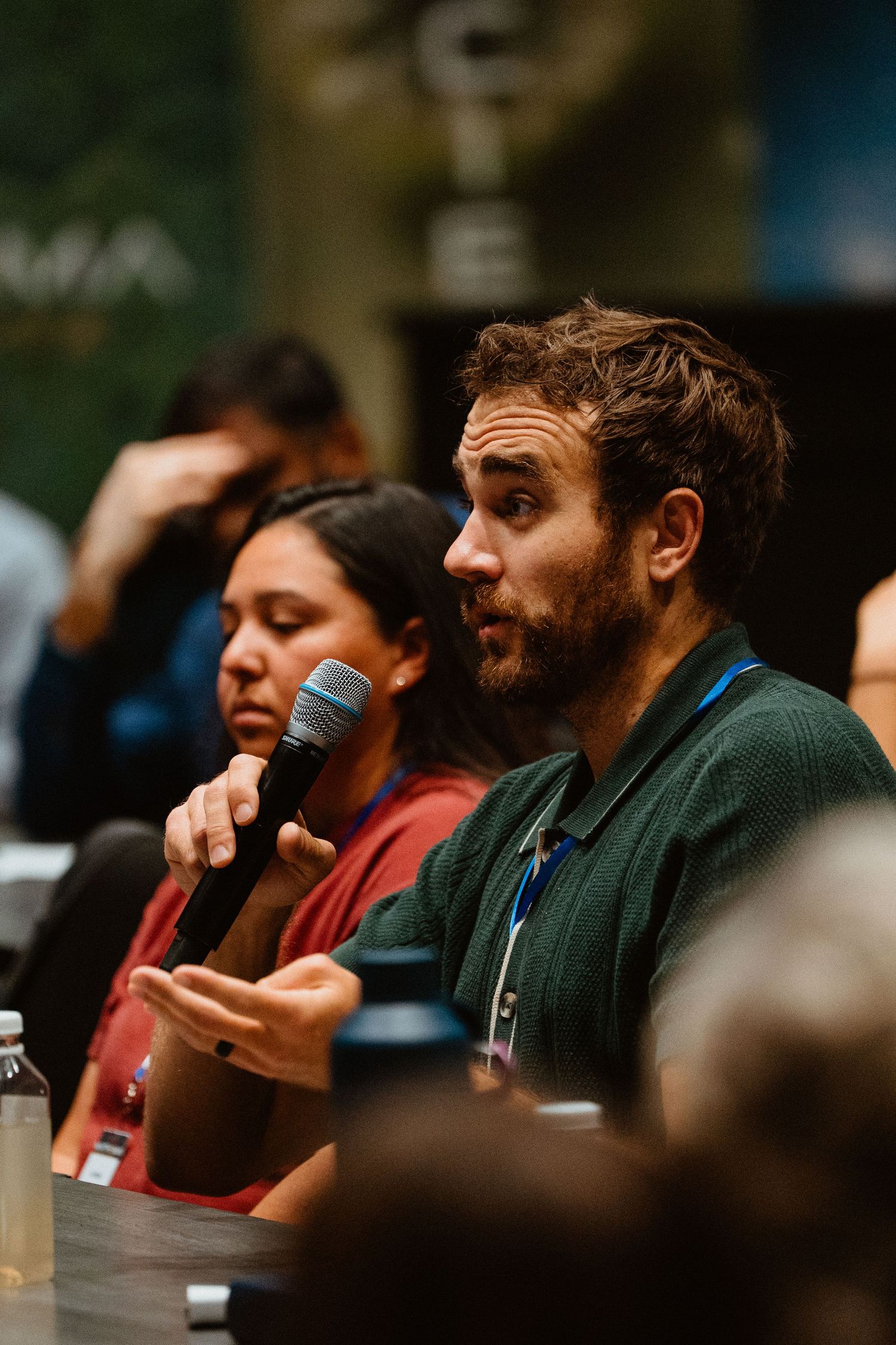 Bearded man speaking into microphone at conference panel, wearing green sweater with blue lanyard.