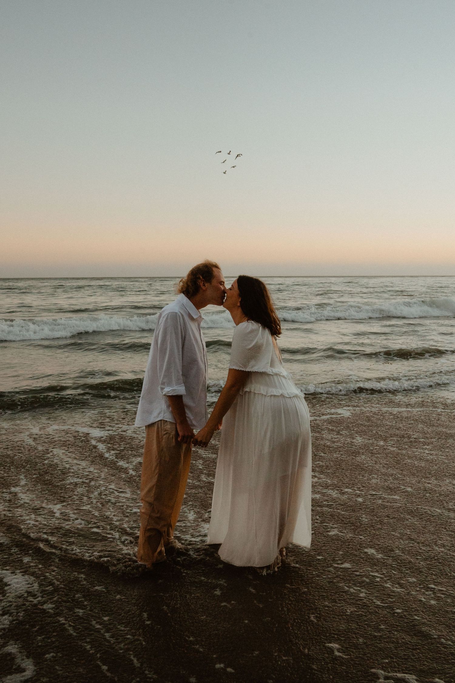 Couple kissing at sunset on a beach, holding hands, woman in white dress, ocean waves in background.