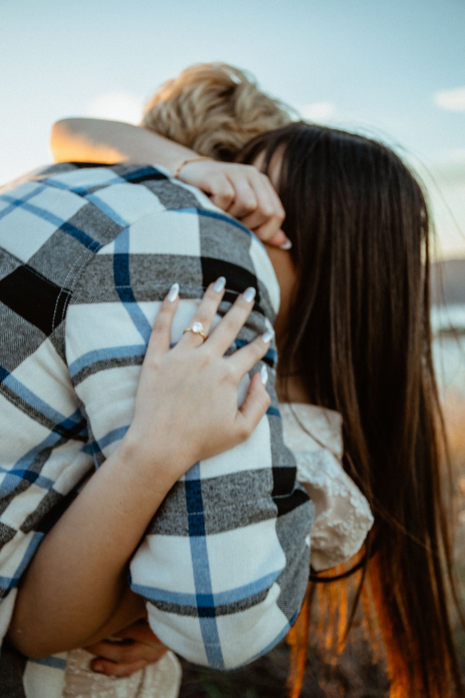 Couple embracing at sunset with woman showing off engagement ring on her hand.