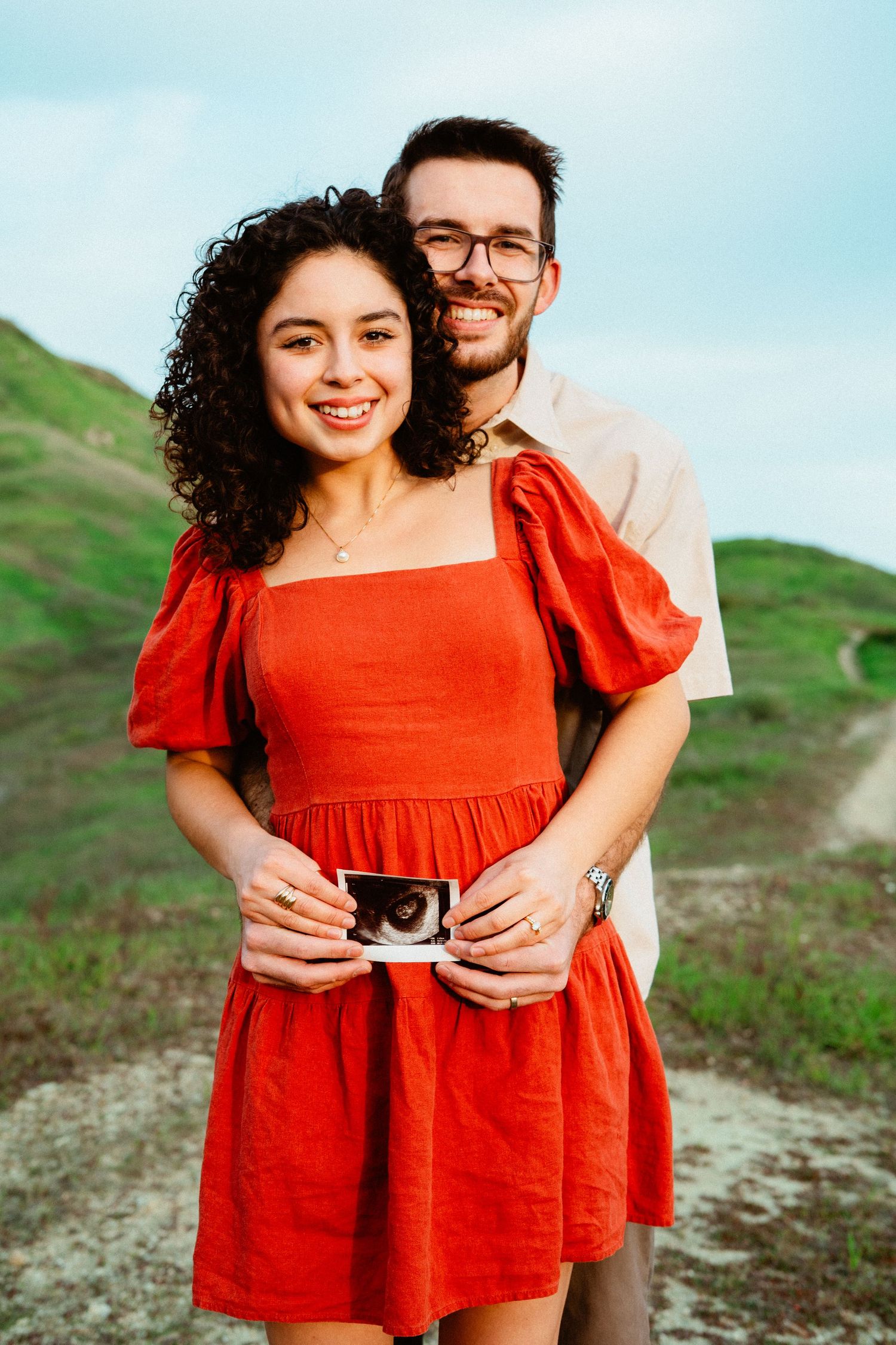 Happy couple holding ultrasound photo outdoors on green hillside, woman in red dress smiling at camera.