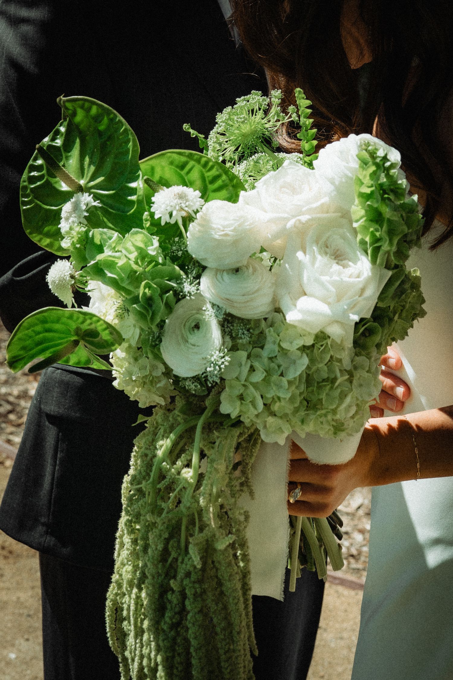 Bride holding lush green and white wedding bouquet with ranunculus, hydrangeas, and tropical leaves.