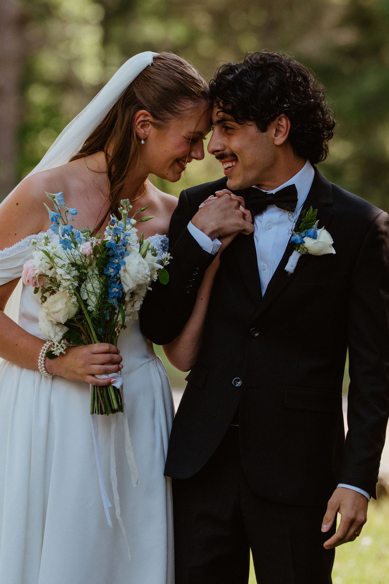 Bride and groom touching foreheads, smiling, she holds blue and white bouquet, he wears black tuxedo with bow tie, outdoors.